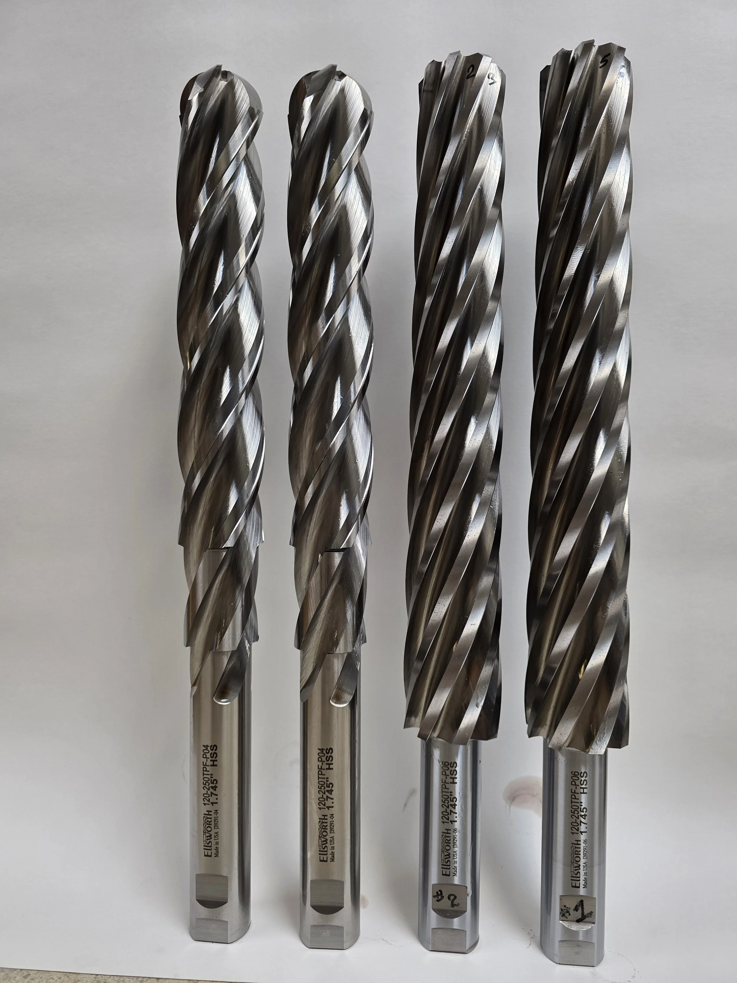 Four large metal drill bits with spiral flutes, standing upright on a plain surface.