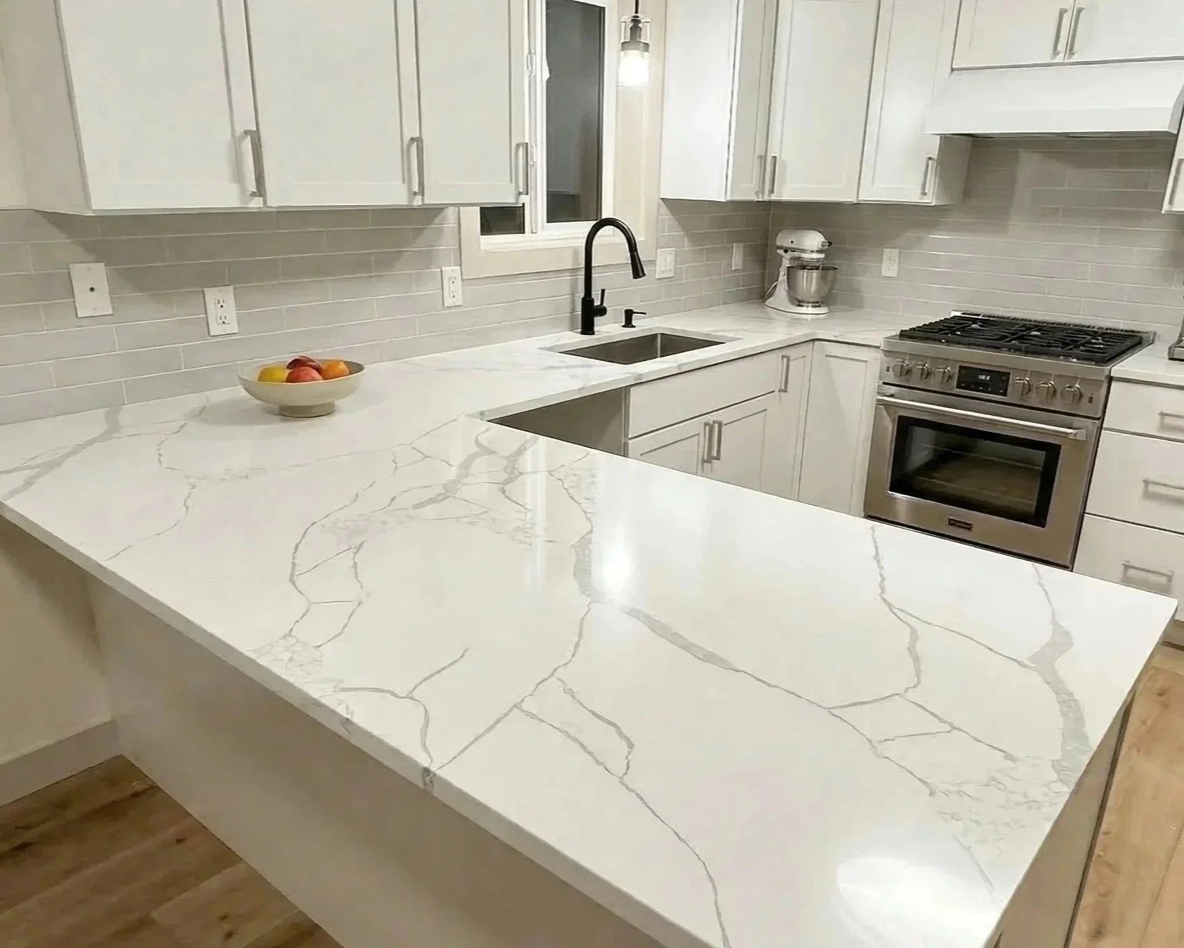 Modern kitchen with white cabinetry, marble countertop with gray veining, stainless steel oven, black faucet, dish soap, bowl of fruit, and light gray tile backsplash.