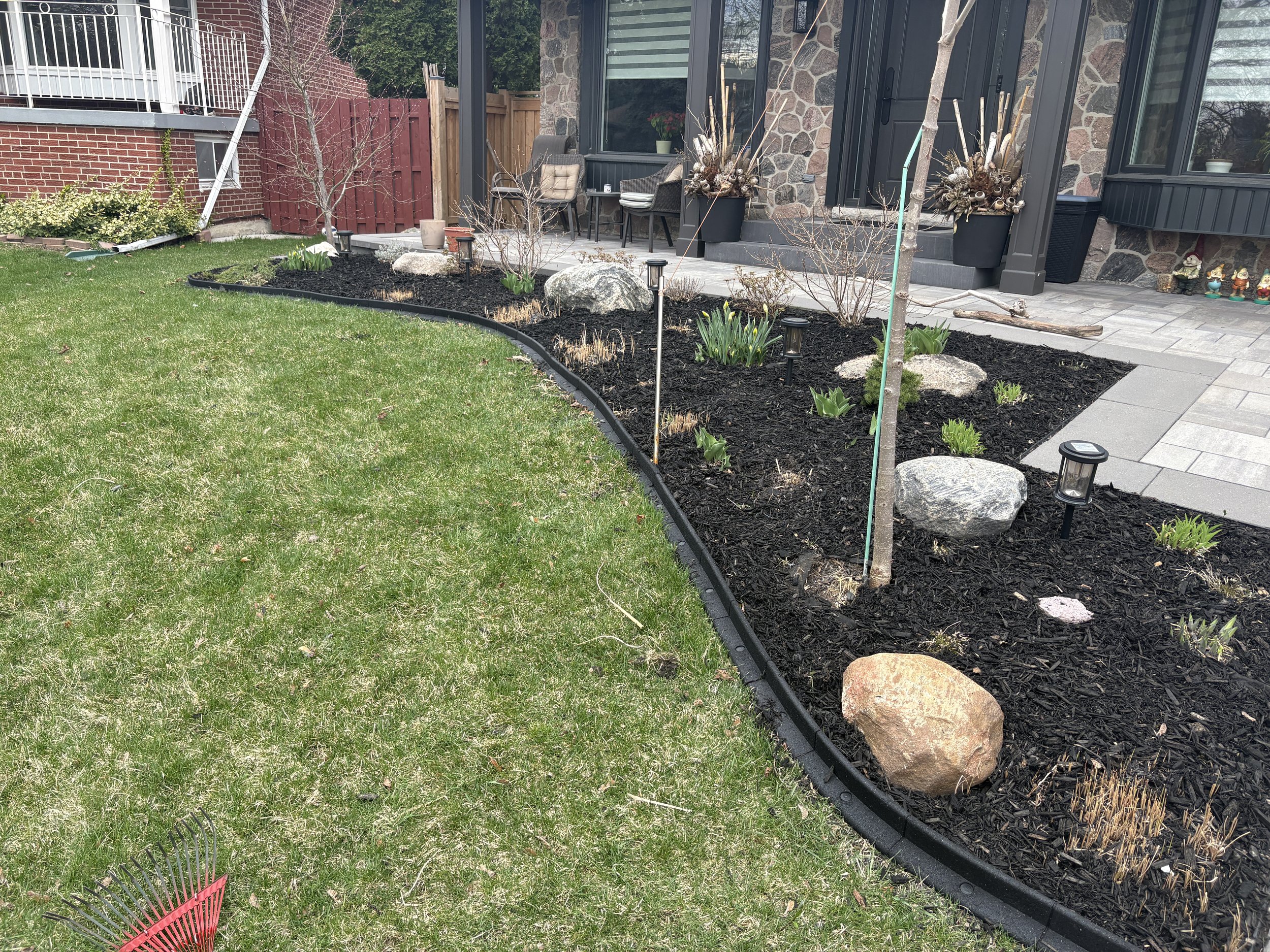A landscaped backyard with a green lawn on the left and a flower bed with dark mulch on the right. The flower bed contains large rocks, small plants, a young tree, and pathway lights. The house has a stone exterior with a porch, window, and door visible.
