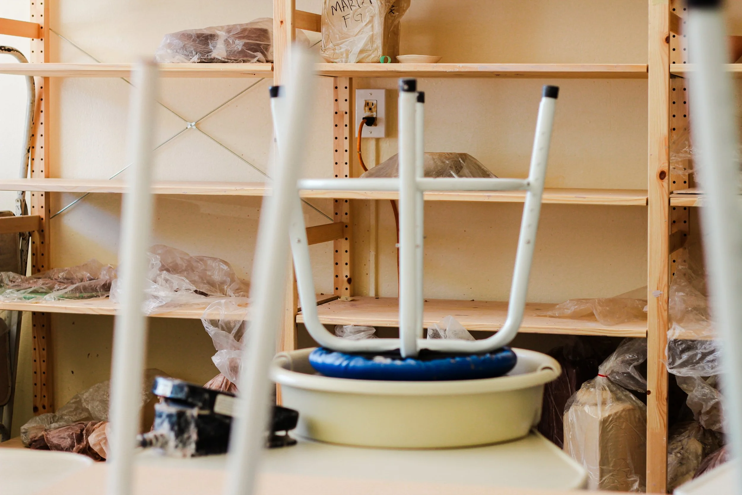 A pottery studio room with wooden shelves holding plastic-wrapped bags of clay. Foreground shows a stool with sitting on a pottery wheel. 