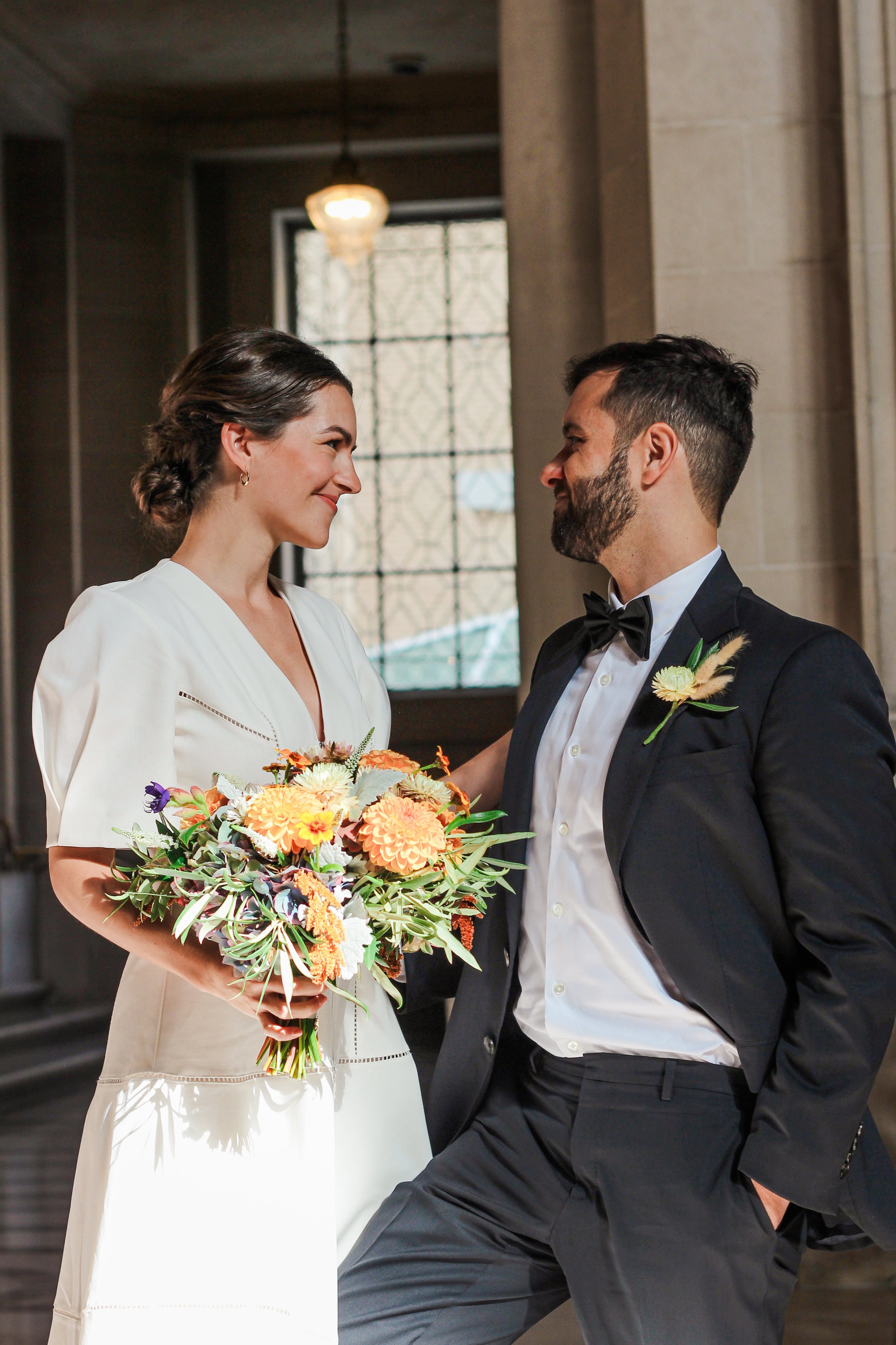 A bride in a white dress and a groom in a black tuxedo with a bow tie standing close inside a historic building with large windows. The bride is holding a colorful bouquet of flowers, and they are smiling at each other.