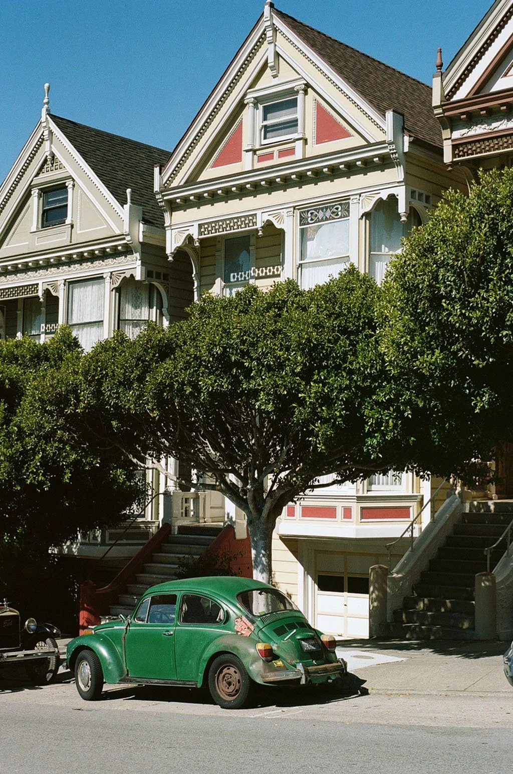 A vintage green Volkswagen Beetle car parked in front of houses with Victorian architecture, trees, and stairs leading up to the houses, under a clear blue sky.