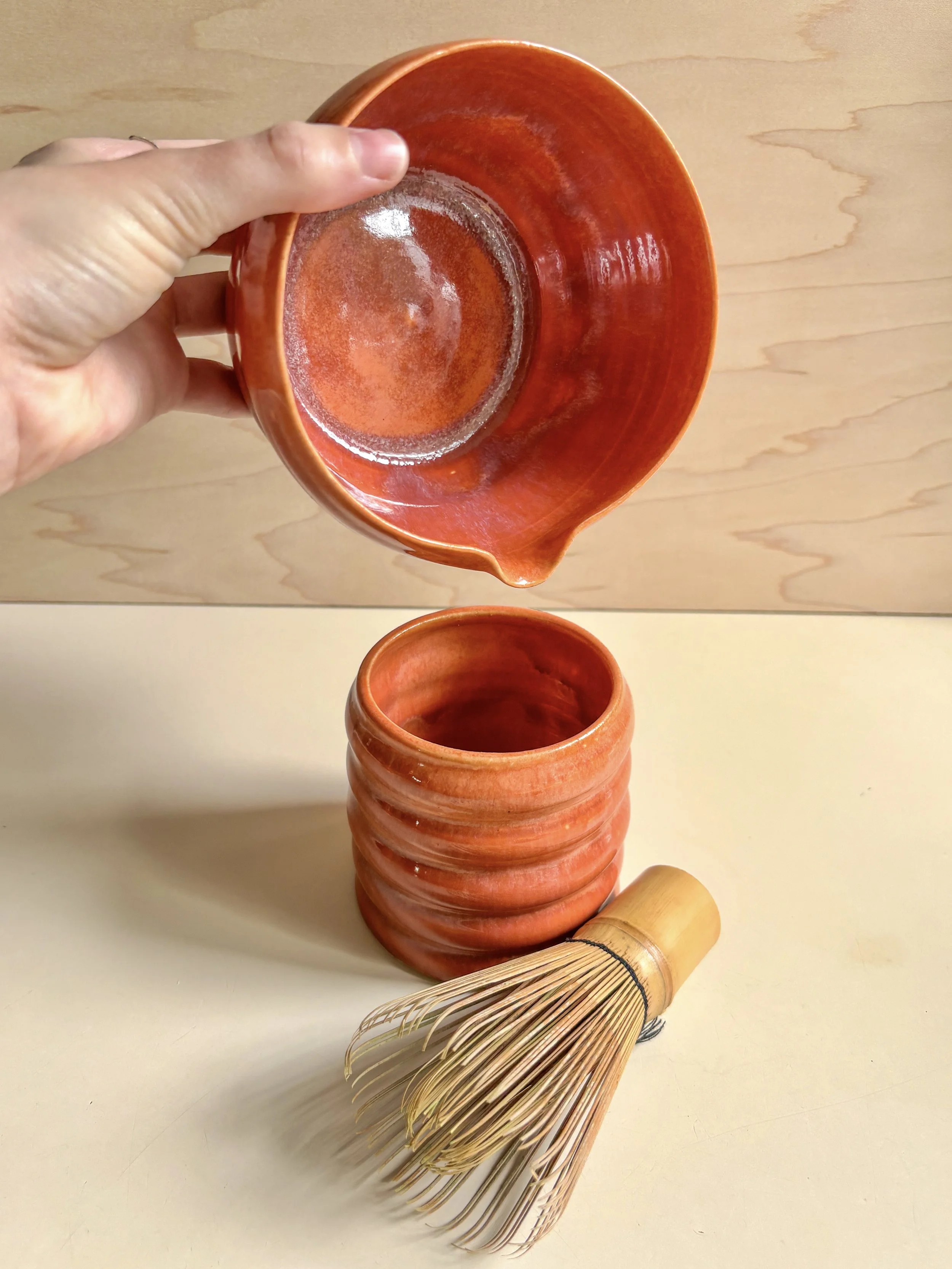 A hand holding a small orange ceramic bowl, a matching orange ceramic cup, and a bamboo whisk resting on a light-colored surface with a wooden background.
