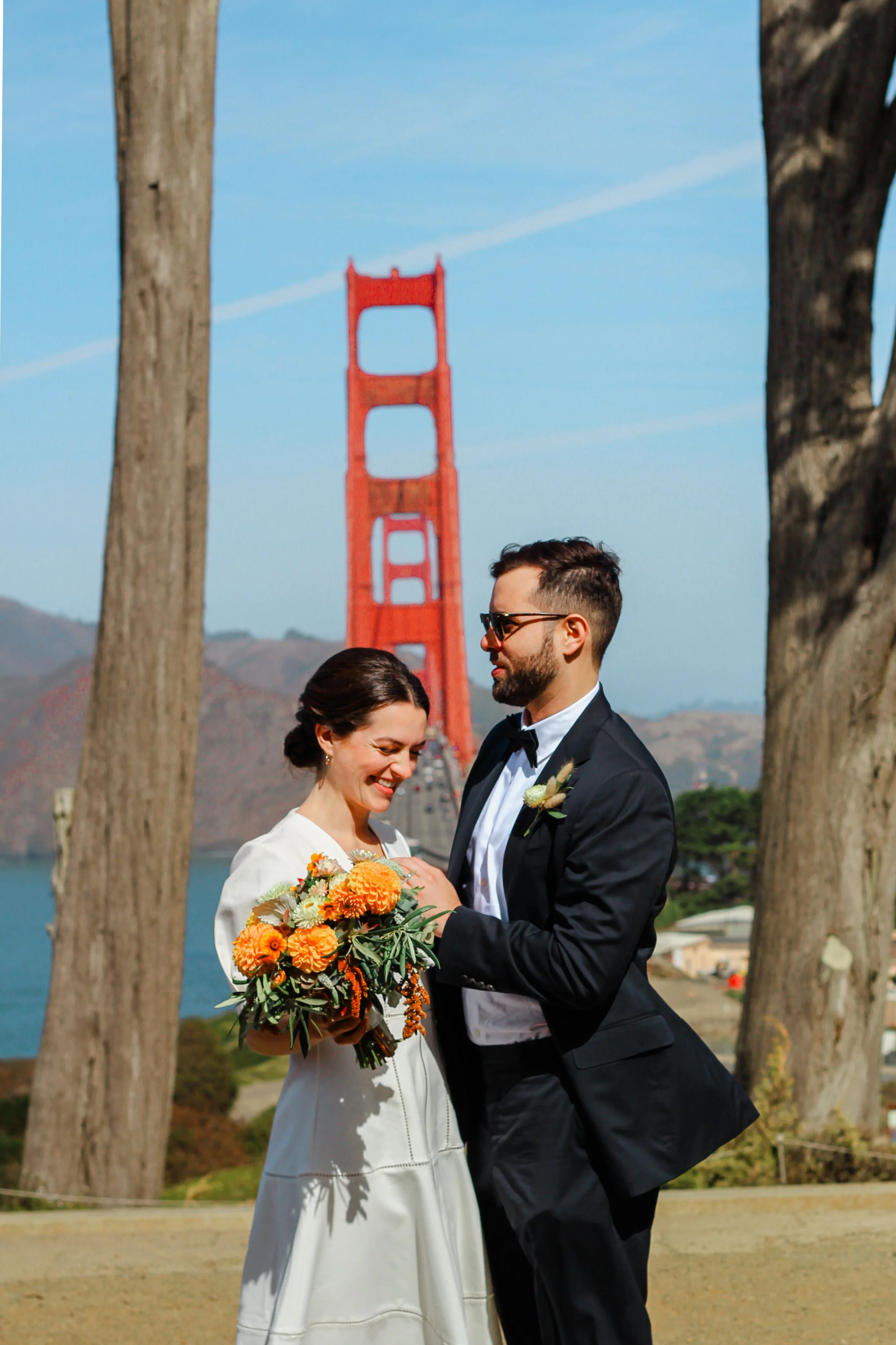 A bride and groom stand together outdoors with the Golden Gate Bridge in the background, surrounded by trees. The bride holds a bouquet of orange and yellow flowers, and both are smiling.