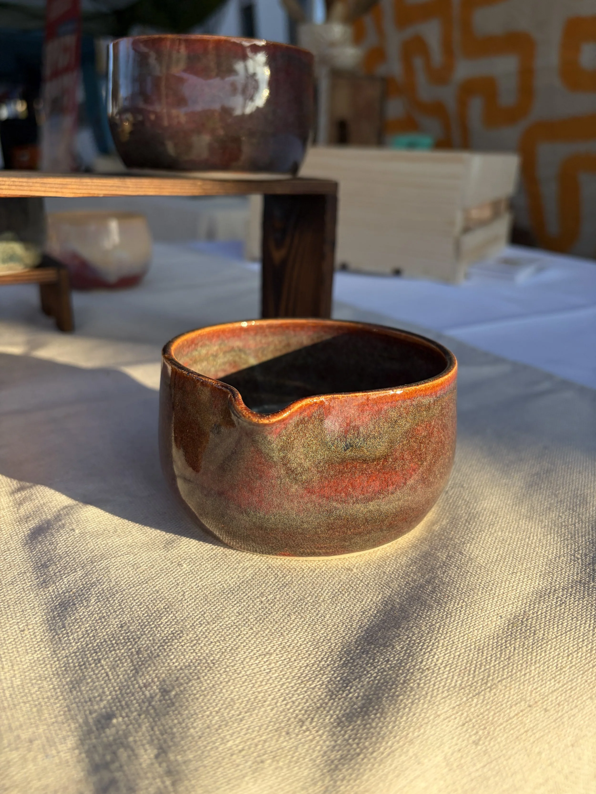 Close-up of a ceramic matcha bowl (chawan) with earthy tones and a small spout, sitting on a table with sunlight casting shadows.