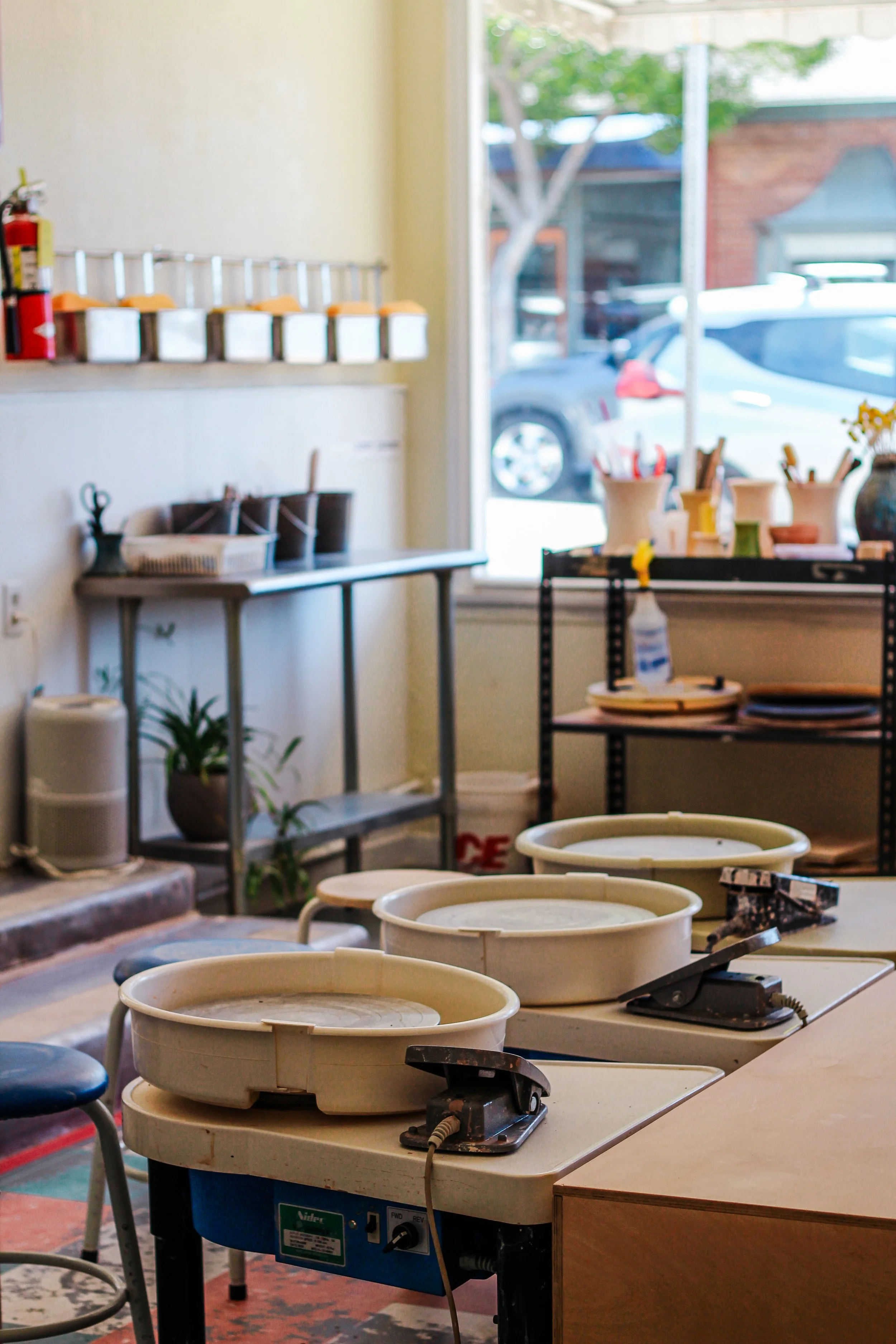 Interior of a pottery studio with pottery wheels on a table and various pottery tools and supplies on shelves near a window.