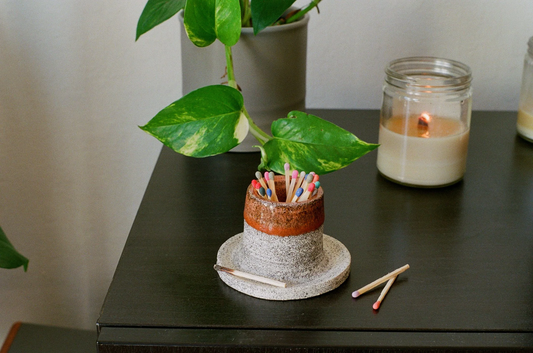 A ceramics match stick holder with colorful matchsticks inside, placed on a round base on a black table. A green leafy plant in a gray pot and candles in glass containers are in the background.