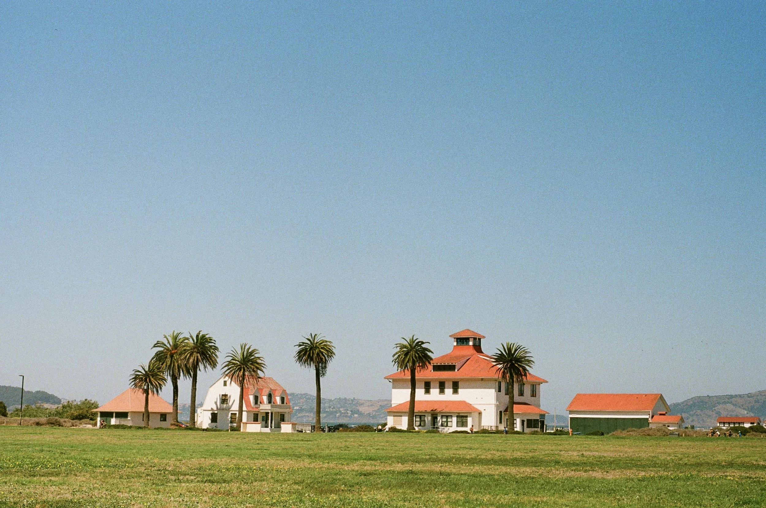 Large house with red-tiled roof, surrounded by tall palm trees, adjacent to smaller buildings, set on a green lawn with distant hills under a clear blue sky.