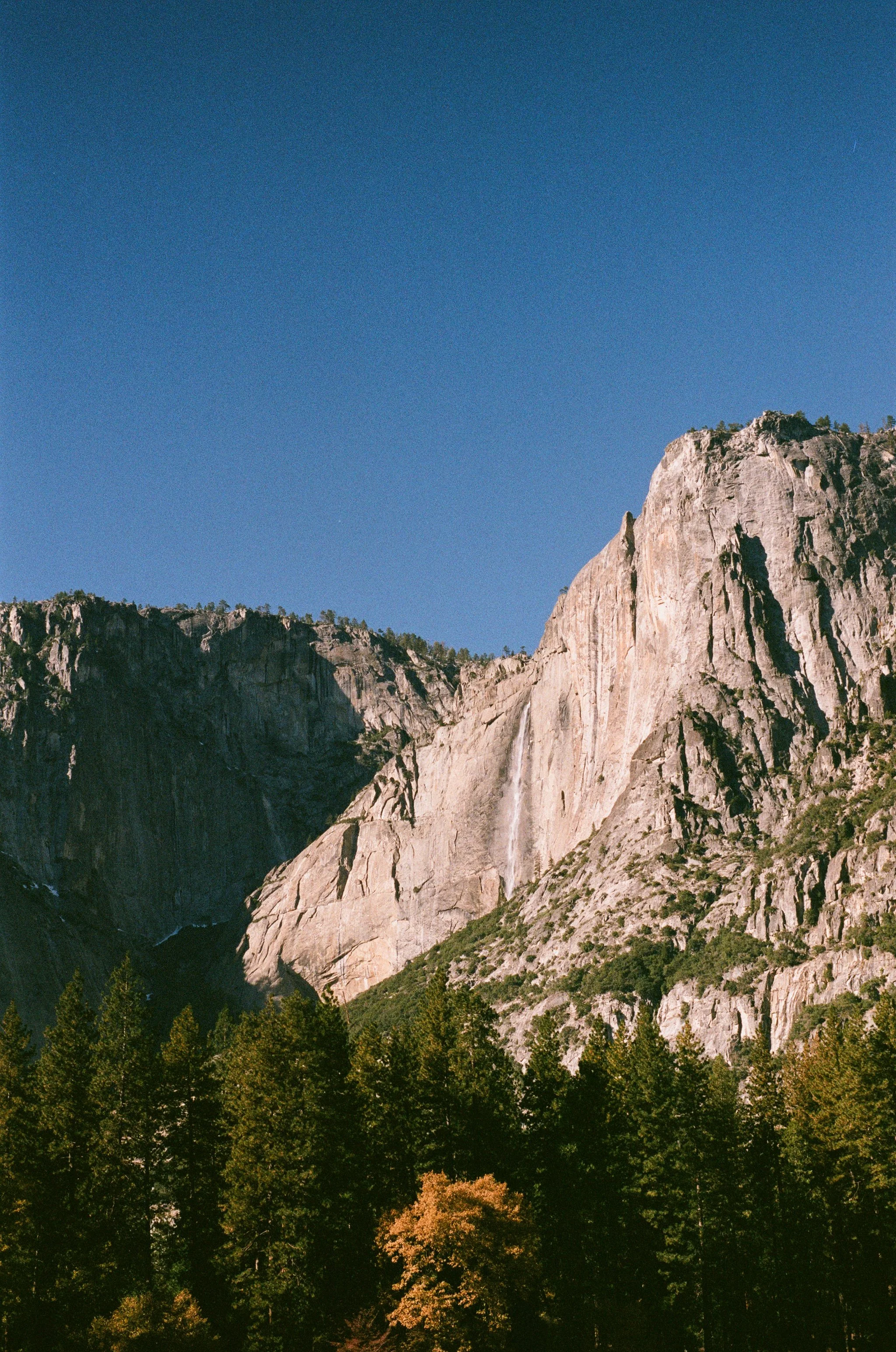 A scenic view of a mountain with a waterfall, trees in the foreground, clear blue sky