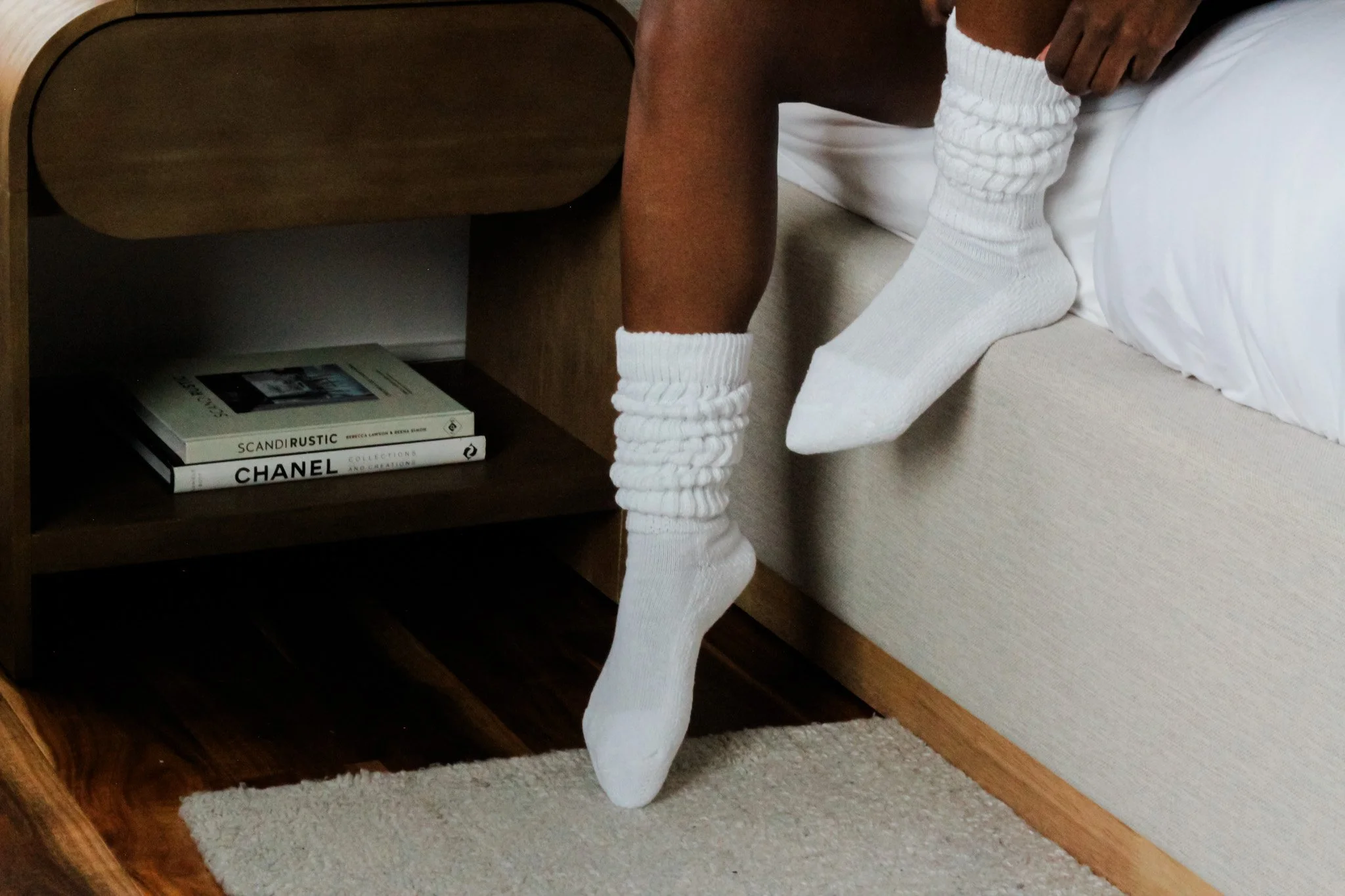 Person sitting on a bed with one leg hanging off, wearing white socks and white shorts, near a wooden nightstand with stacked books.