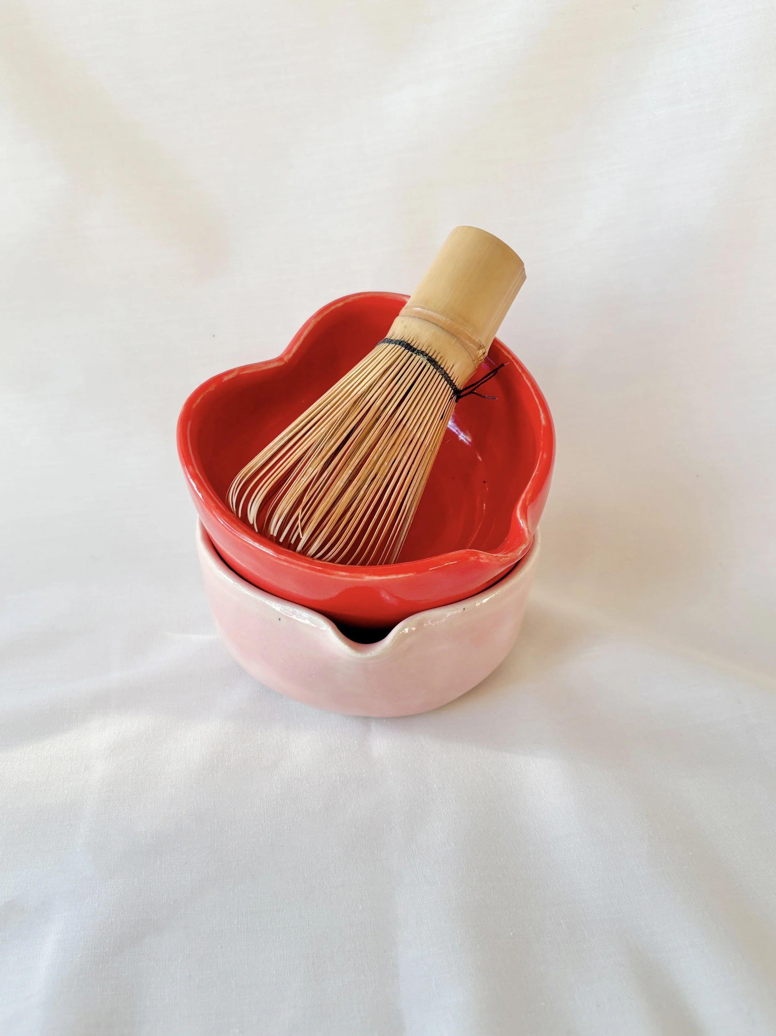 A bamboo matcha whisk resting inside a red ceramic heart-shaped matcha bowl (Chawan), with a pink bowl beneath it on a white surface.