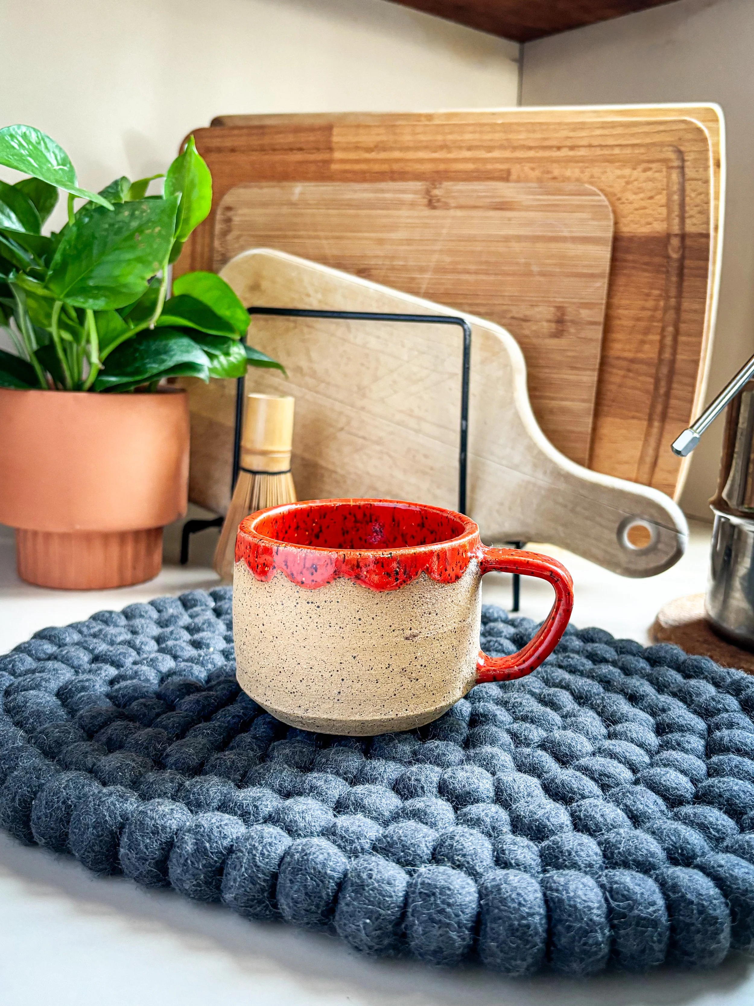 A ceramic mug with raw speckled buff clay body and red glazed rim and handle sitting on a textured gray felt mat. Behind the mug, there are several wooden cutting boards, a potted plant, and a small bamboo whisk on a kitchen counter.