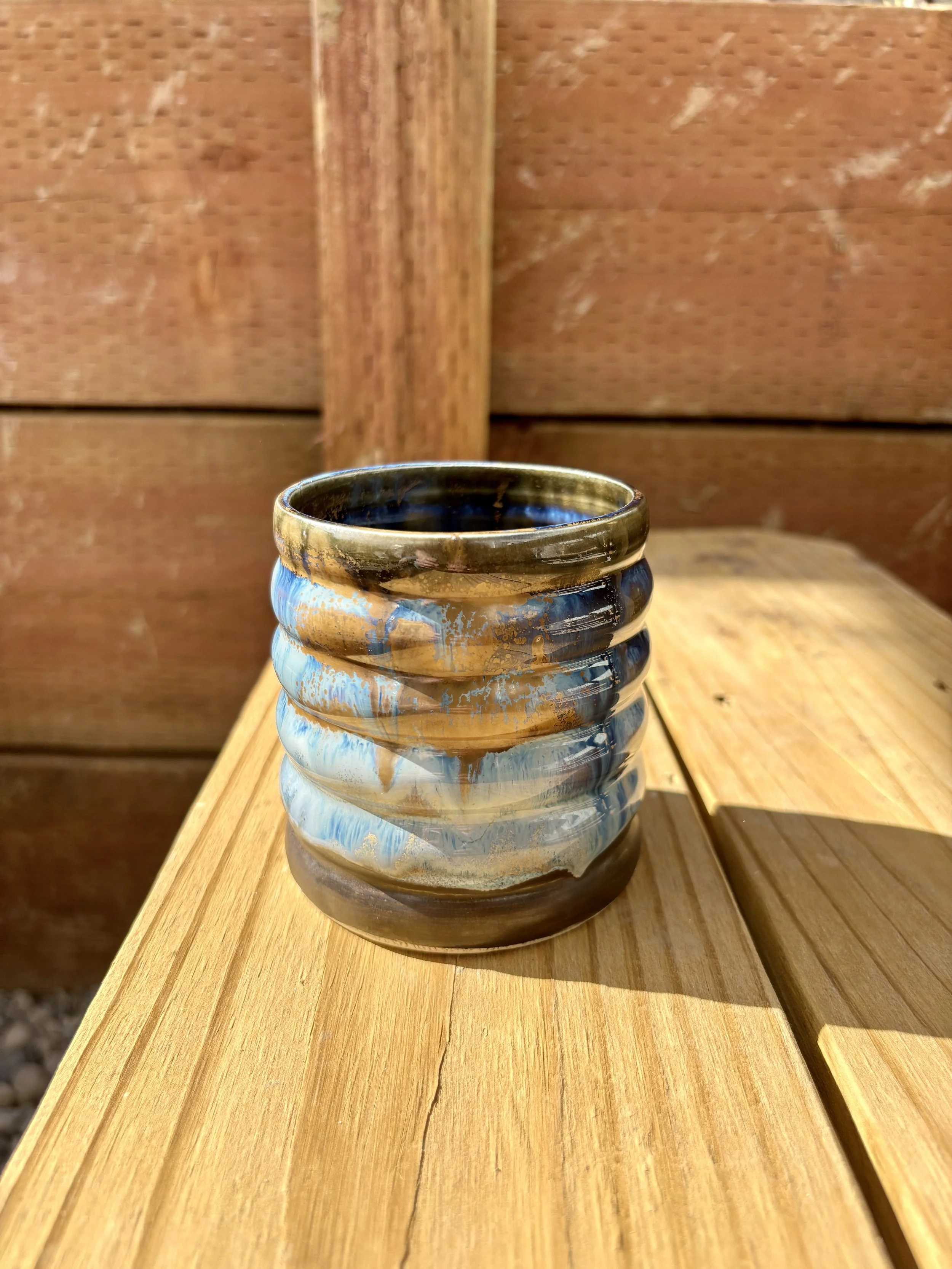 A ceramic cup with a ribbed texture and swirling blue and gold glaze, sitting on a wooden surface outdoors with a wooden fence in the background.