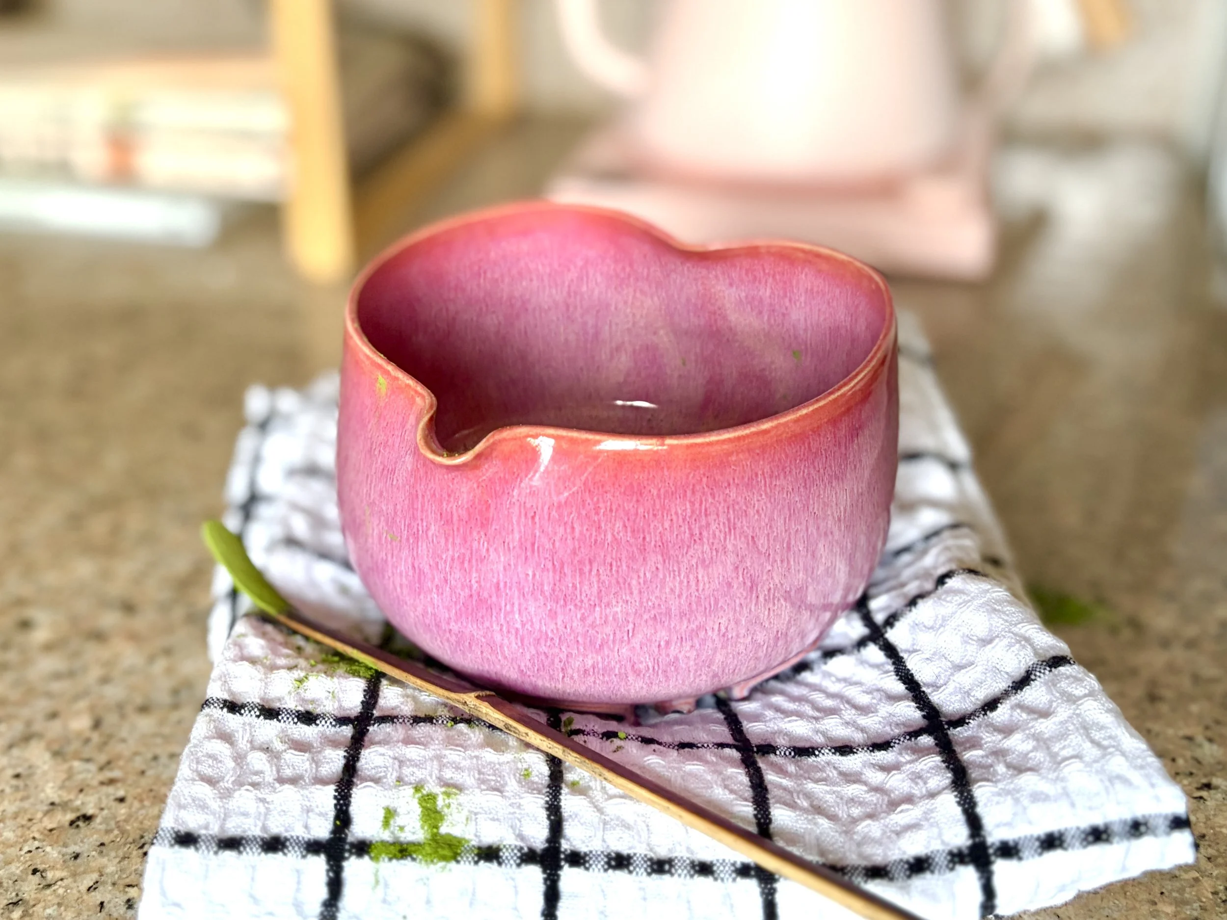 A pink, heart-shaped ceramic matcha bowl (chawan), sitting on a black and white checkered cloth on a kitchen countertop, with a wooden spoon and matcha powder nearby.