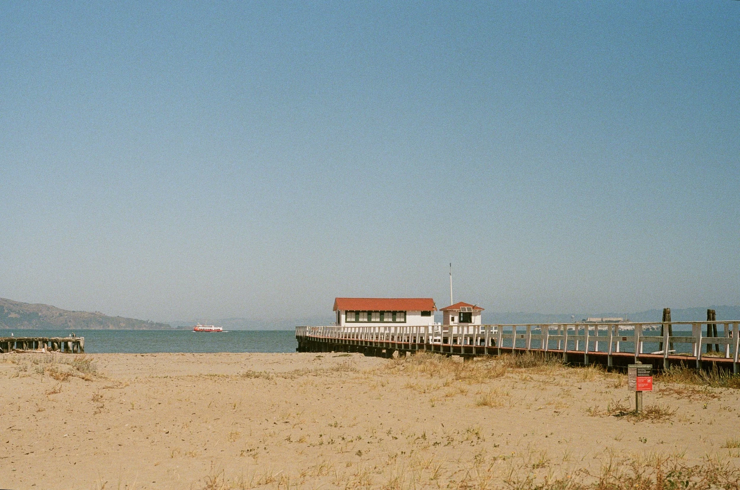 A sandy beach leading to a pier with a small building with red roof, overlooking the water with a ship in the distance, under a clear blue sky.