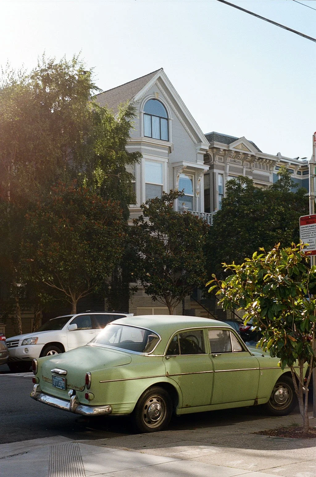 A vintage green car parked on the street with modern houses and trees in the background.