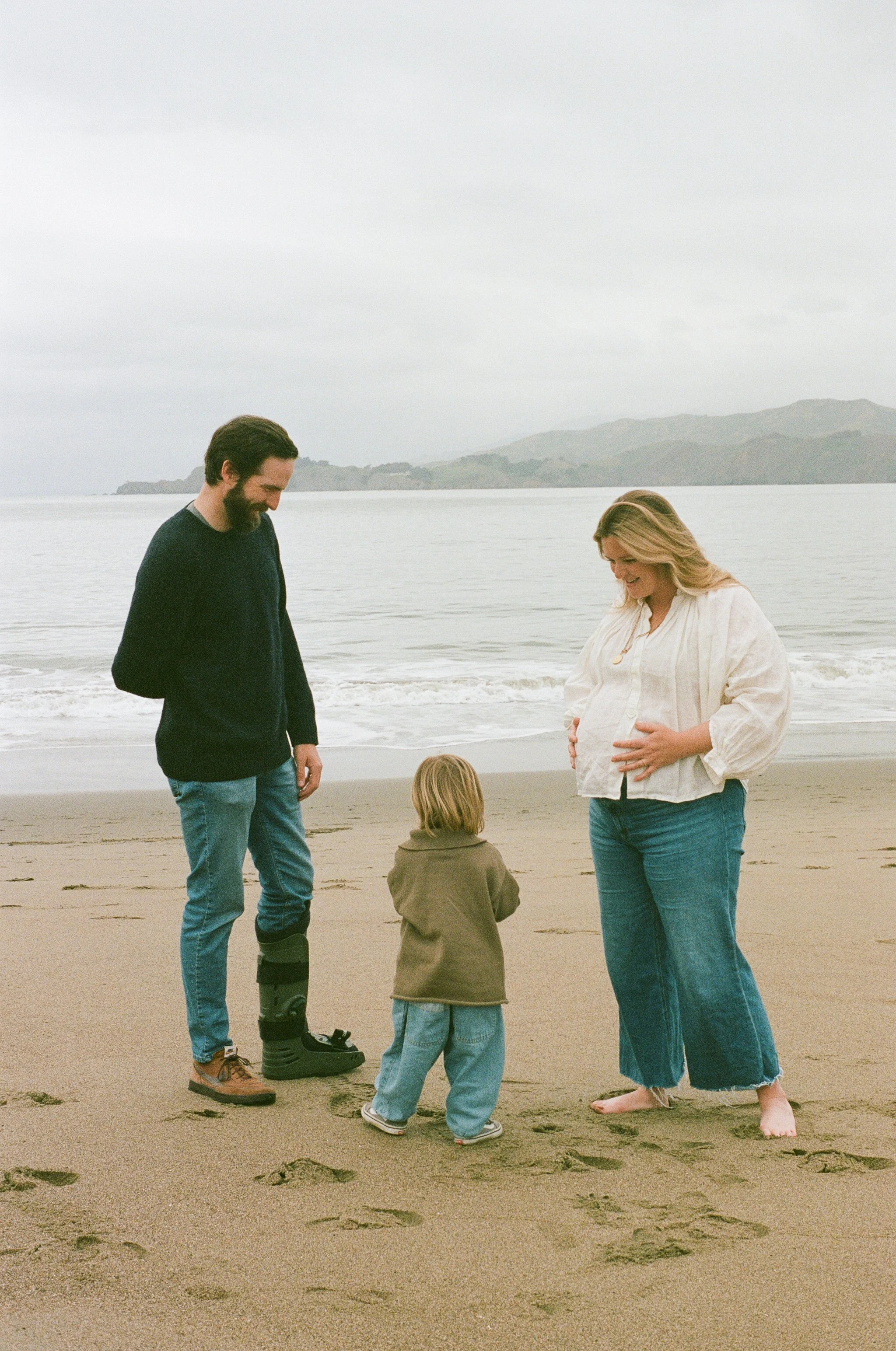 A family of three on a beach, with a pregnant woman, a man, and a young child standing together near the shoreline, smiling and having a moment.