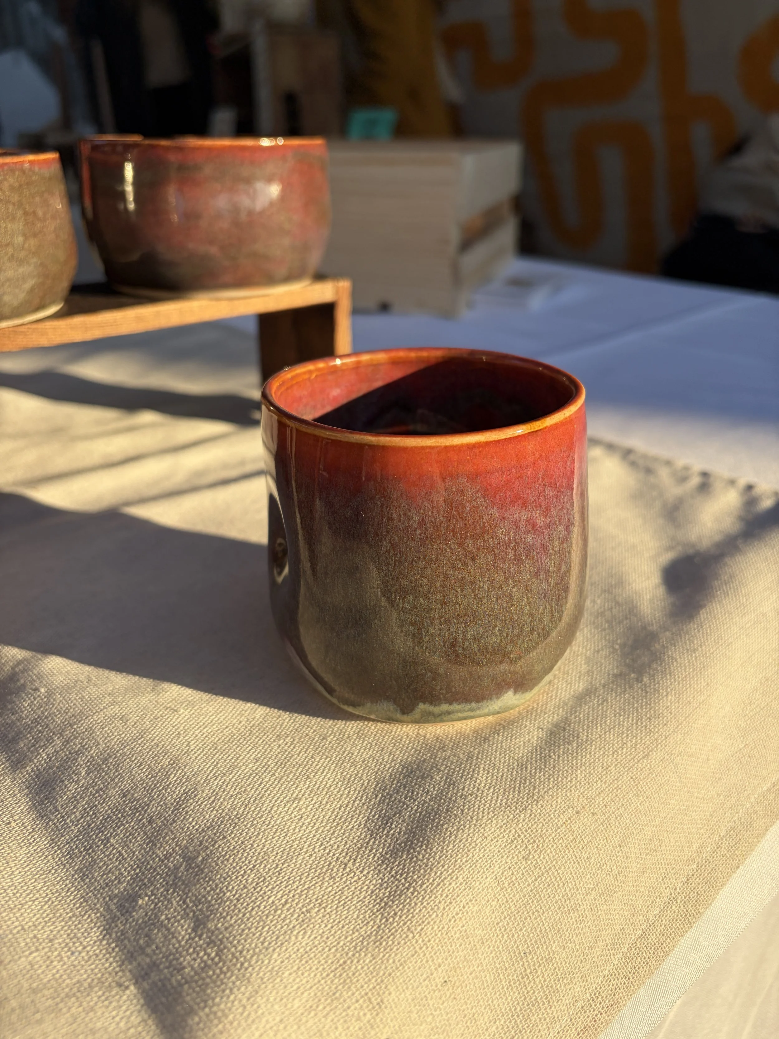 Close-up of a red and purple glazed ceramic cup on a beige tablecloth with shadows, with wooden shelves and more cups in the background.