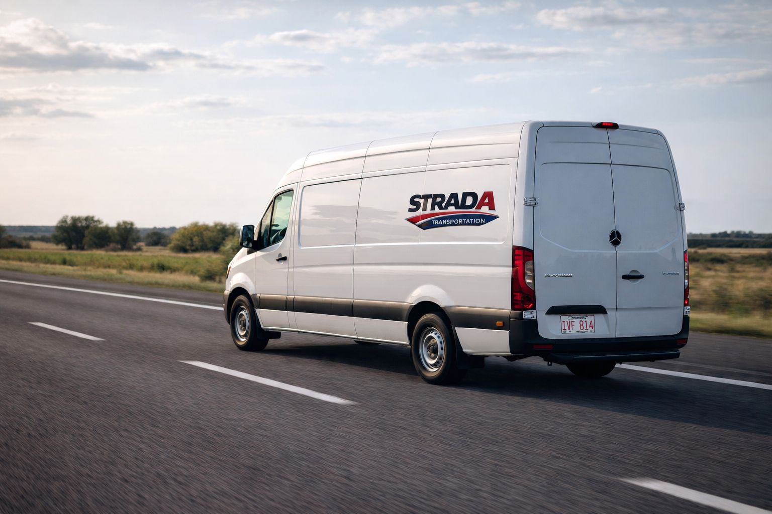 White cargo van with 'STRADA TRANSPORTATION' logo driving on a highway with grassy fields and trees in the background under a partly cloudy sky.