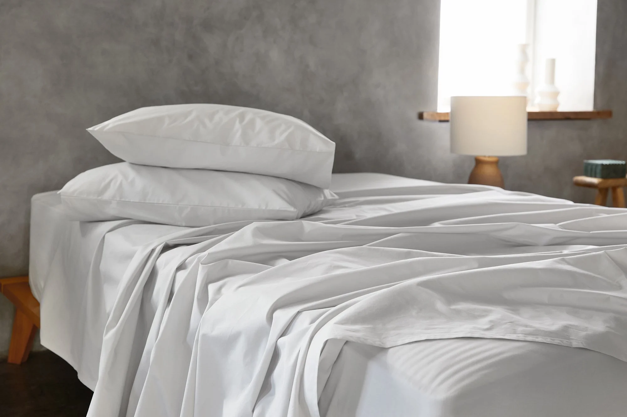 A neatly made bed with white bed sheets and two pillows against a gray wall. There is a wooden bedside table with a ceramic cup, a lamp with a wooden base, and a window with a wooden sill and white vases on it.