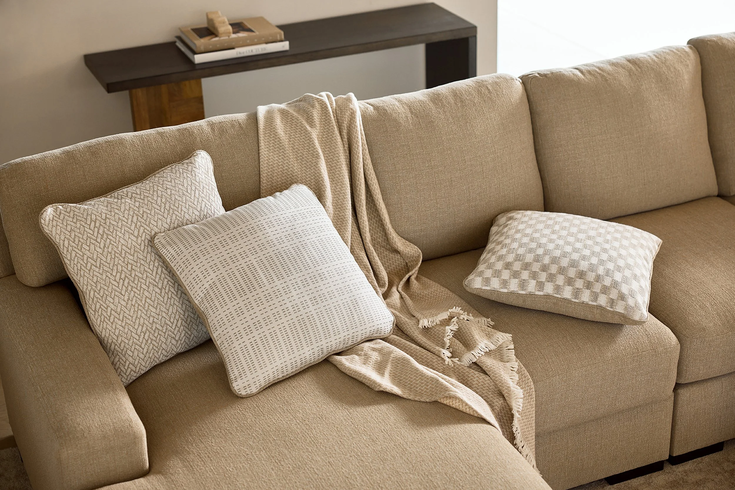 A beige sofa with a beige throw blanket, three patterned throw pillows, and a beige blanket draped over the backrest. In the background, there is a dark wooden console table with a stack of books and a small decorative object on top.