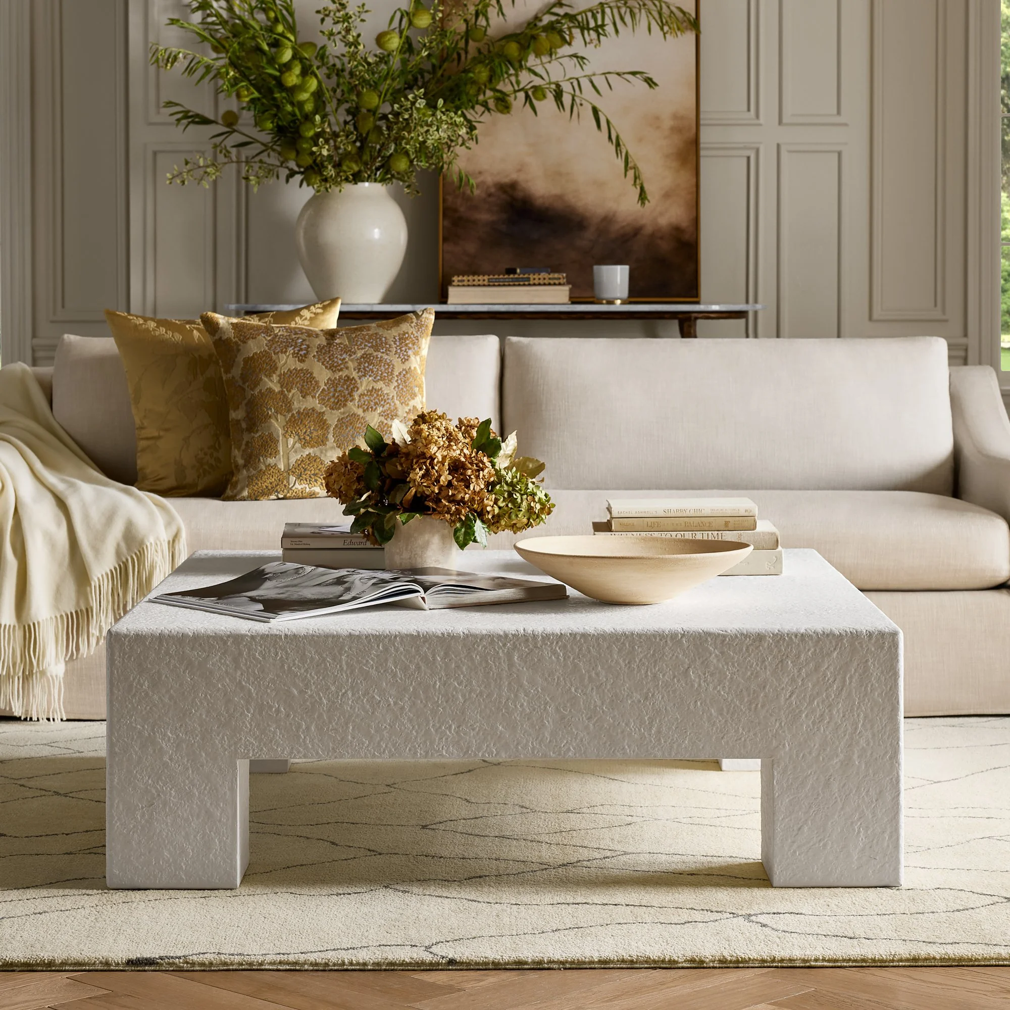 A living room with a cream-colored sofa with gold floral pillows, a coffee table with books and a flower arrangement, a textured white side table, and a painting on the wall behind a large white vase with green foliage. The room has paneled walls and