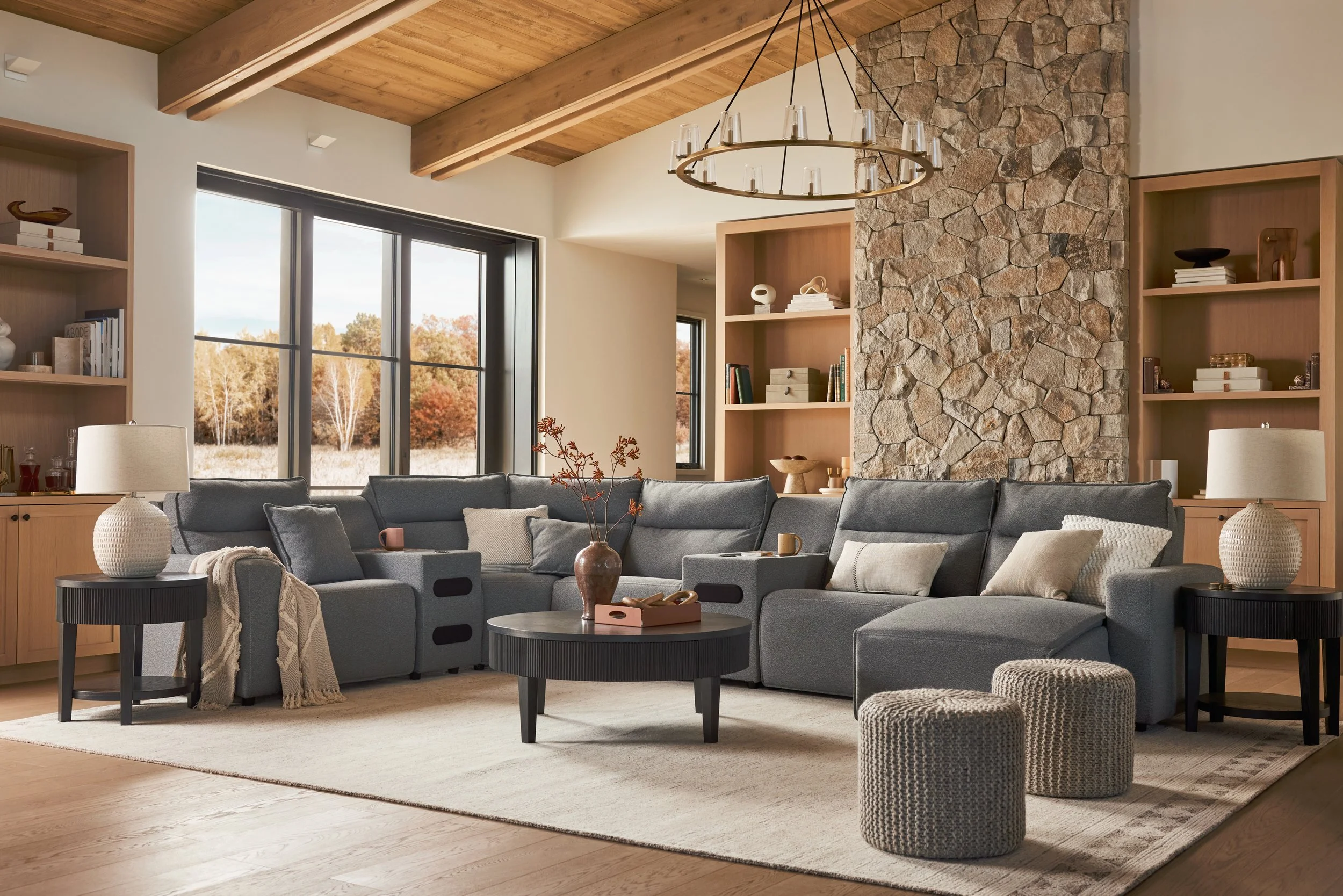 Living room with a gray sectional sofa, round black coffee tables, beige rug, and textured poufs. Large windows reveal trees outside with fall foliage. Wooden bookshelves, stone accent wall, and a chandelier hang from a wooden ceiling.