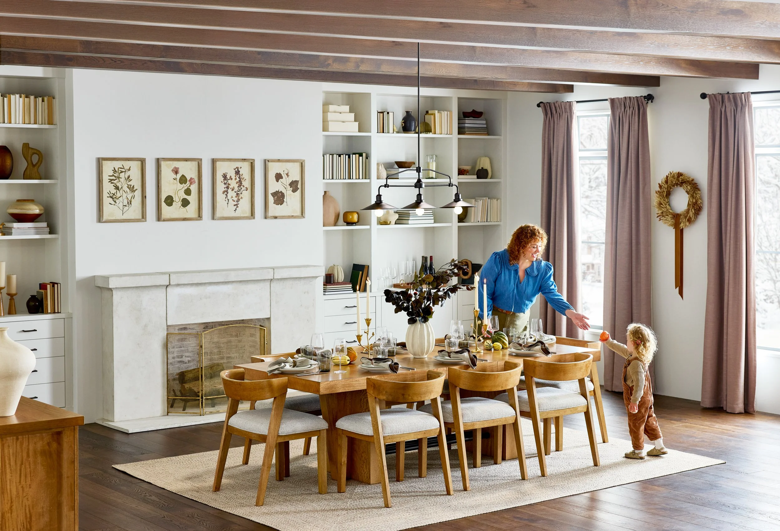 A cozy dining room with a wooden table set for a meal, surrounded by wooden chairs. A woman and a young girl are enjoying their time together, the woman reaching across the table, and the girl holding a small orange object. The room features a white 