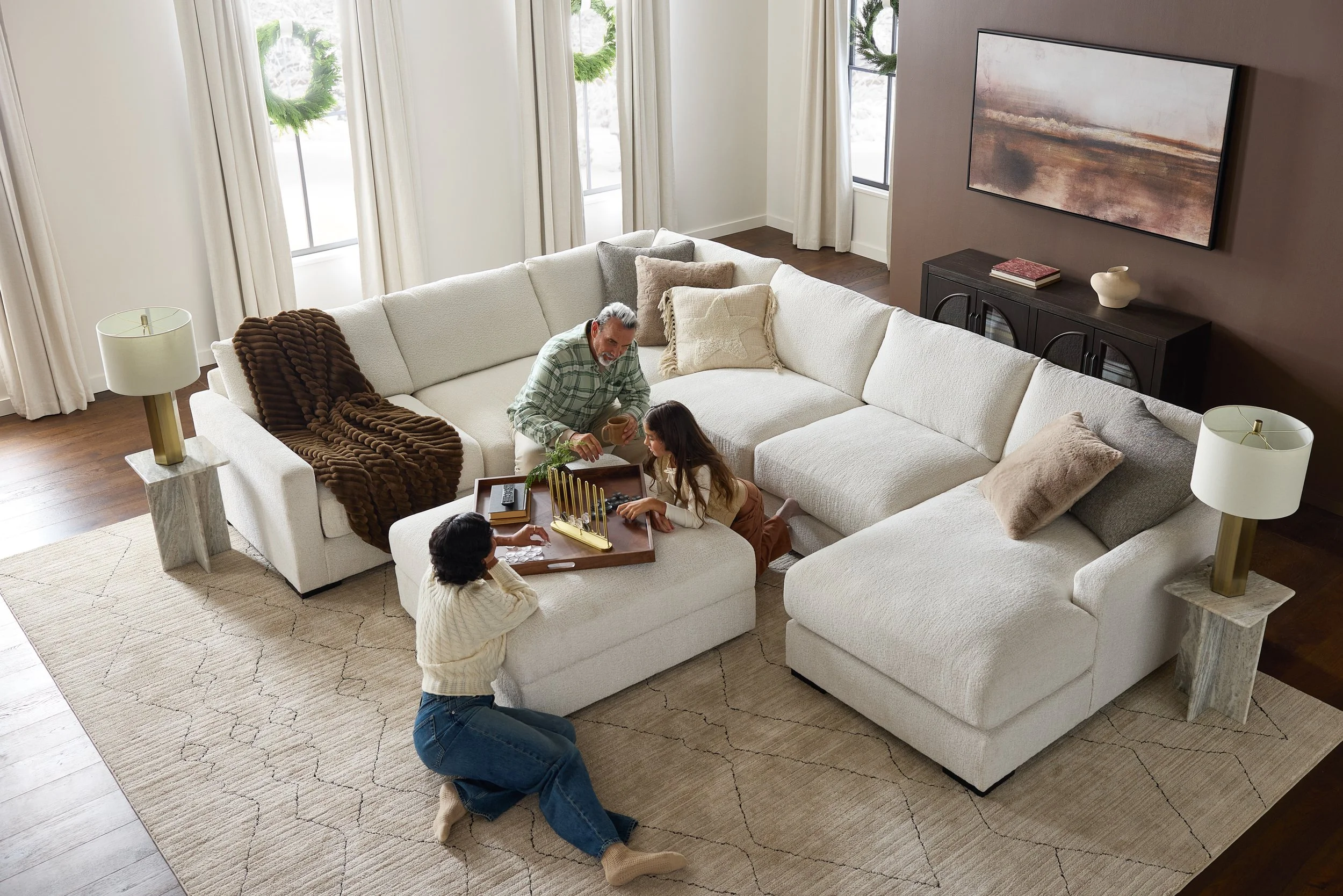 A living room with a large white sectional sofa and three people playing a game around a table. The room has cream-colored curtains, a large abstract painting on a brown wall, and two white table lamps with gold bases.