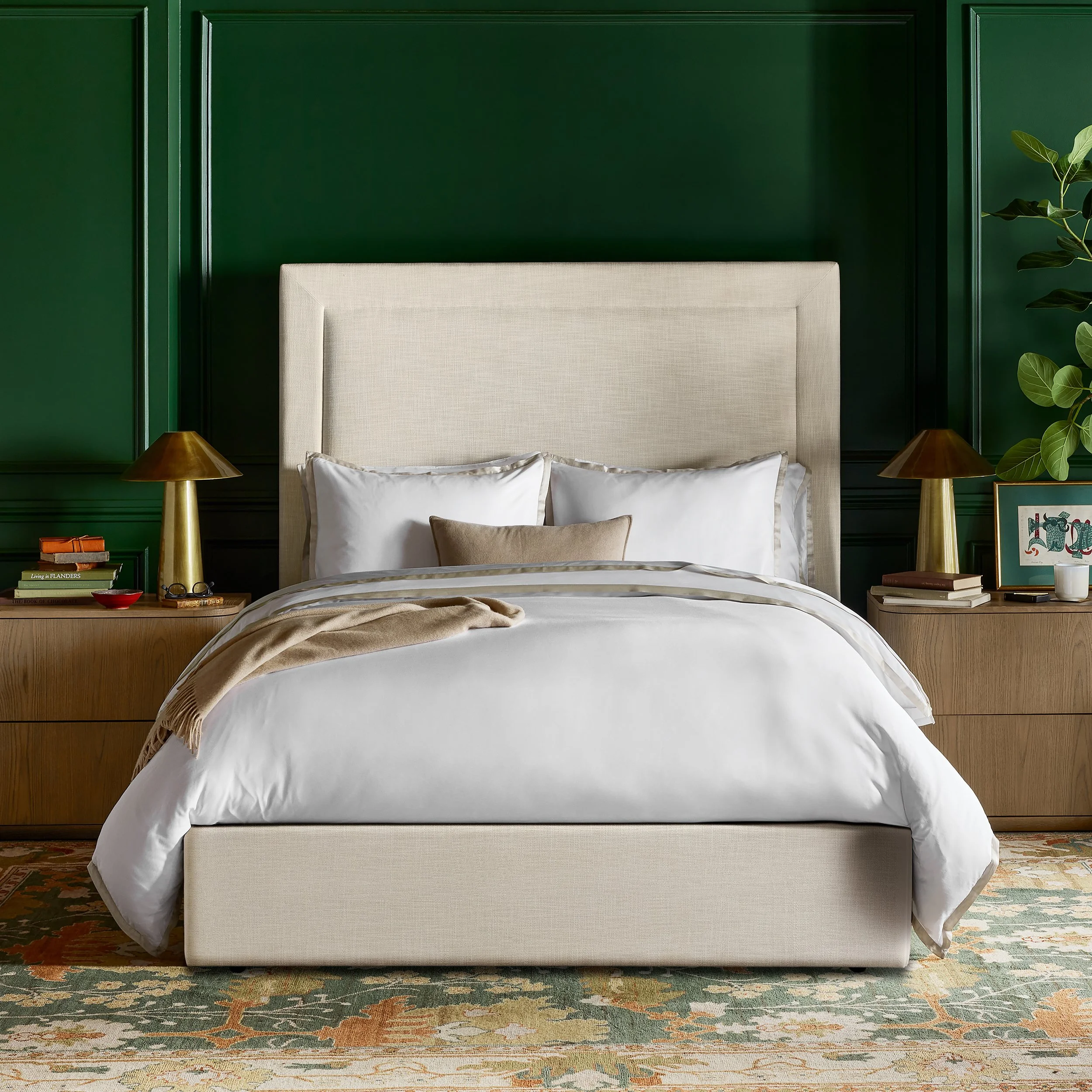 A bedroom with a beige upholstered bed with white bedding and a beige throw blanket. On each side of the bed are wooden nightstands with brass lamps. There are stacks of books and decorative items on the nightstands. The dark green wall behind the be