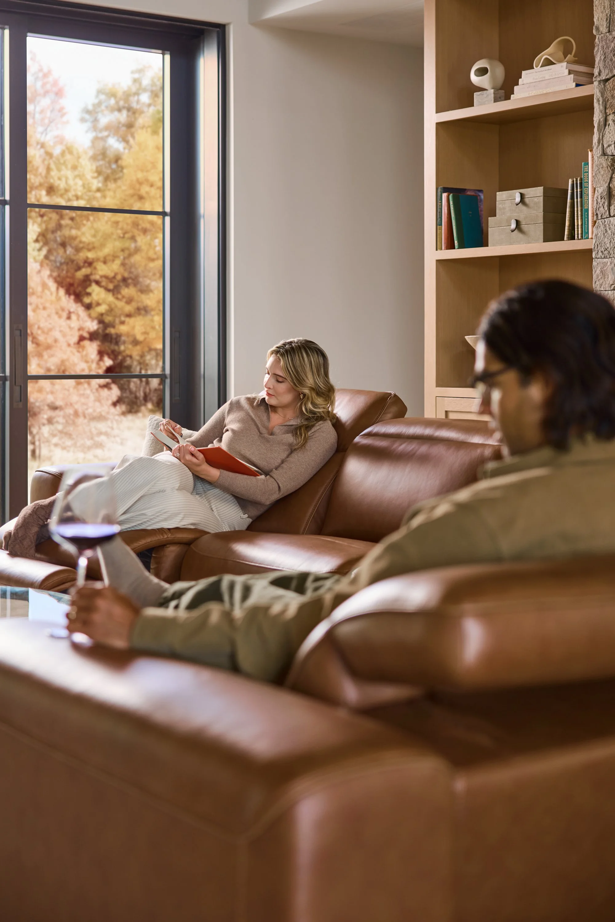 Two women sitting on a leather sofa in a living room with wooden shelves and a window with autumn trees outside.