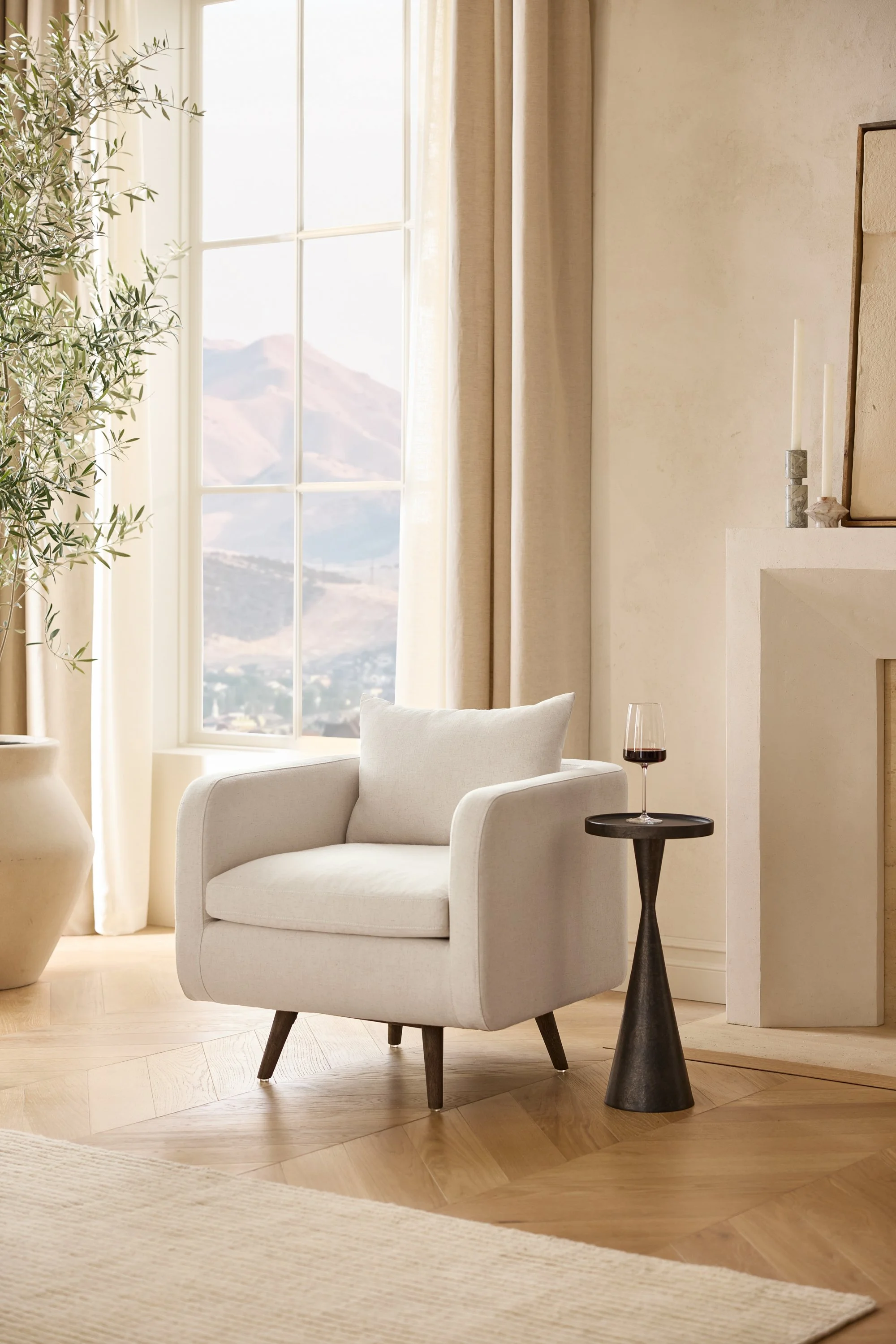A cozy living room with a white armchair, a small black side table with a glass of red wine, a large window with mountain views, beige curtains, and a beige fireplace mantel with candles and decorative items.