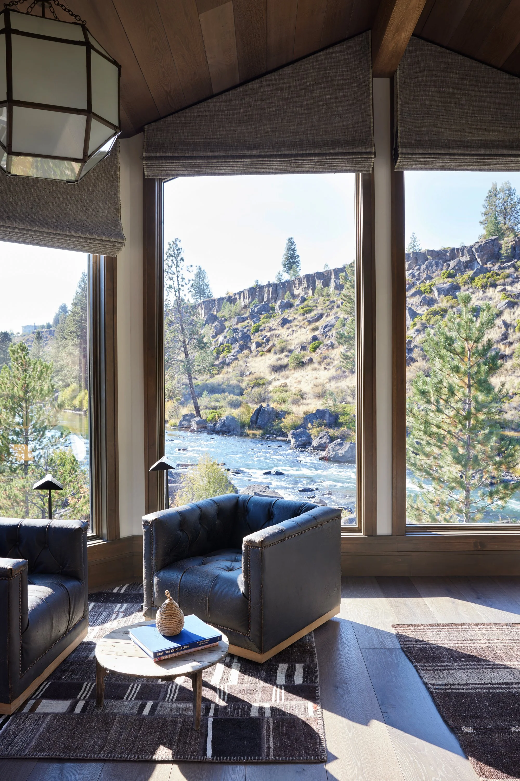 Living room with large windows showing a view of trees, rocks, and a river outside. Inside, there are two black leather armchairs, a small round marble-topped table with books and a decorative object, and a geometric pendant light. The room has wood-