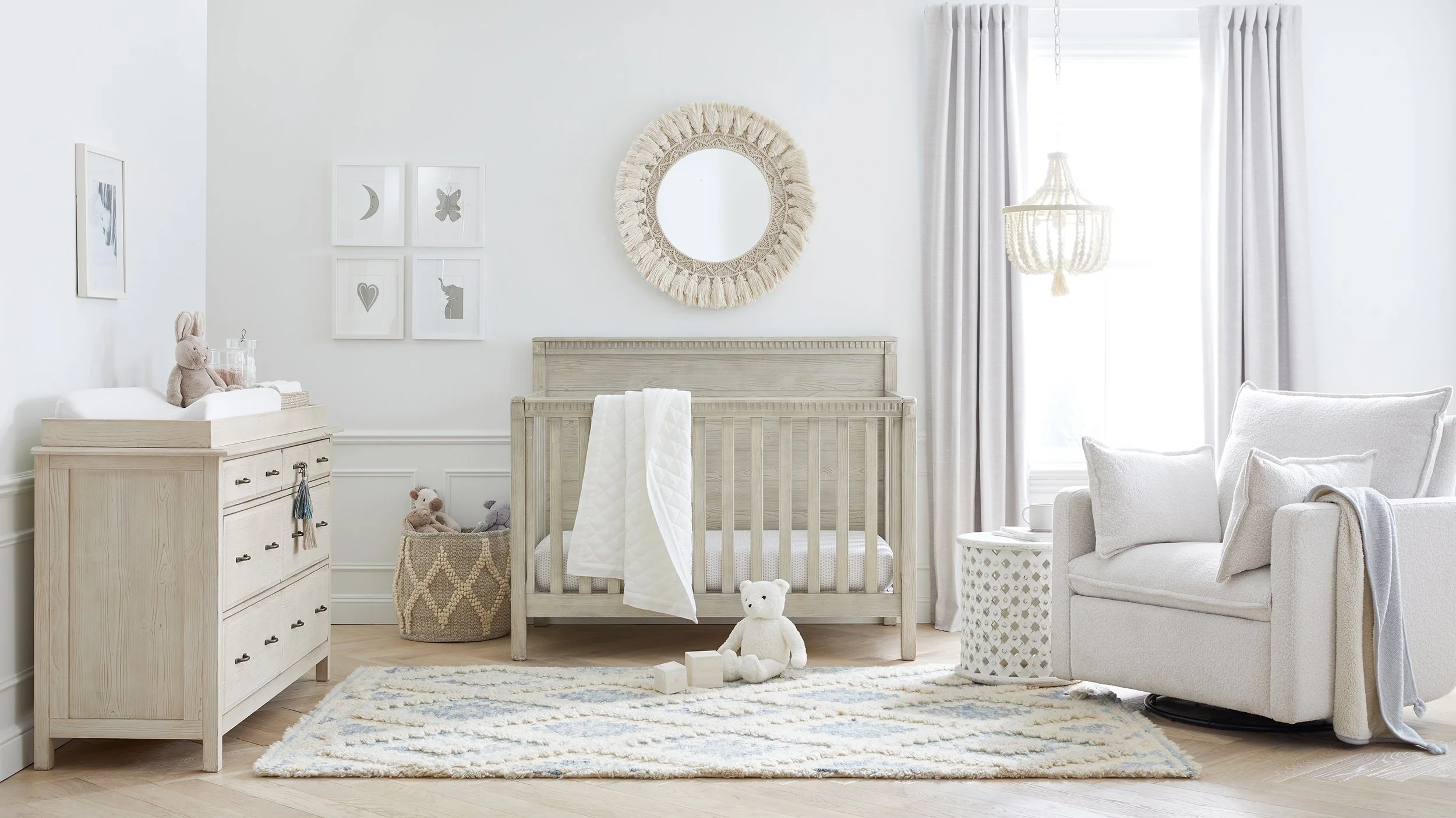 A nursery room with white walls, a wooden crib with a white blanket, a white armchair with pillows, a dresser with toys, framed moon and butterfly wall art, a round woven mirror, curtains, a chandelier, and stuffed animals.