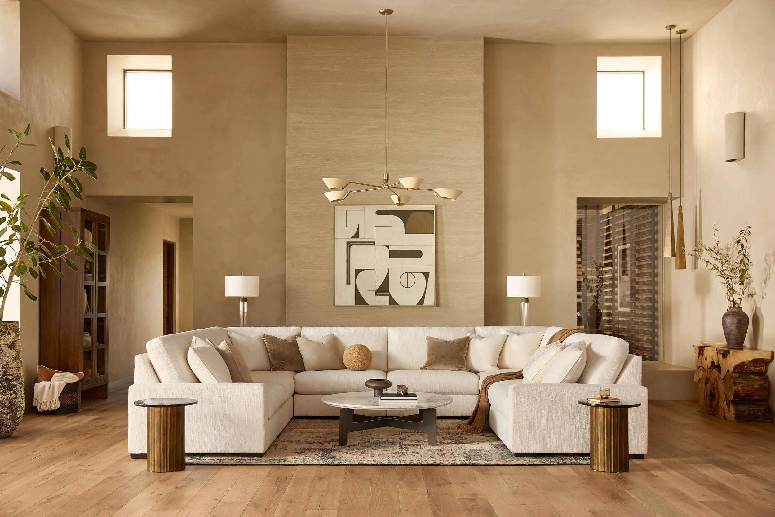 A modern living room with a beige sofa, decorative pillows, a round coffee table, and artistic wall decor. Large windows, plants, and elegant lighting create a cozy ambiance.