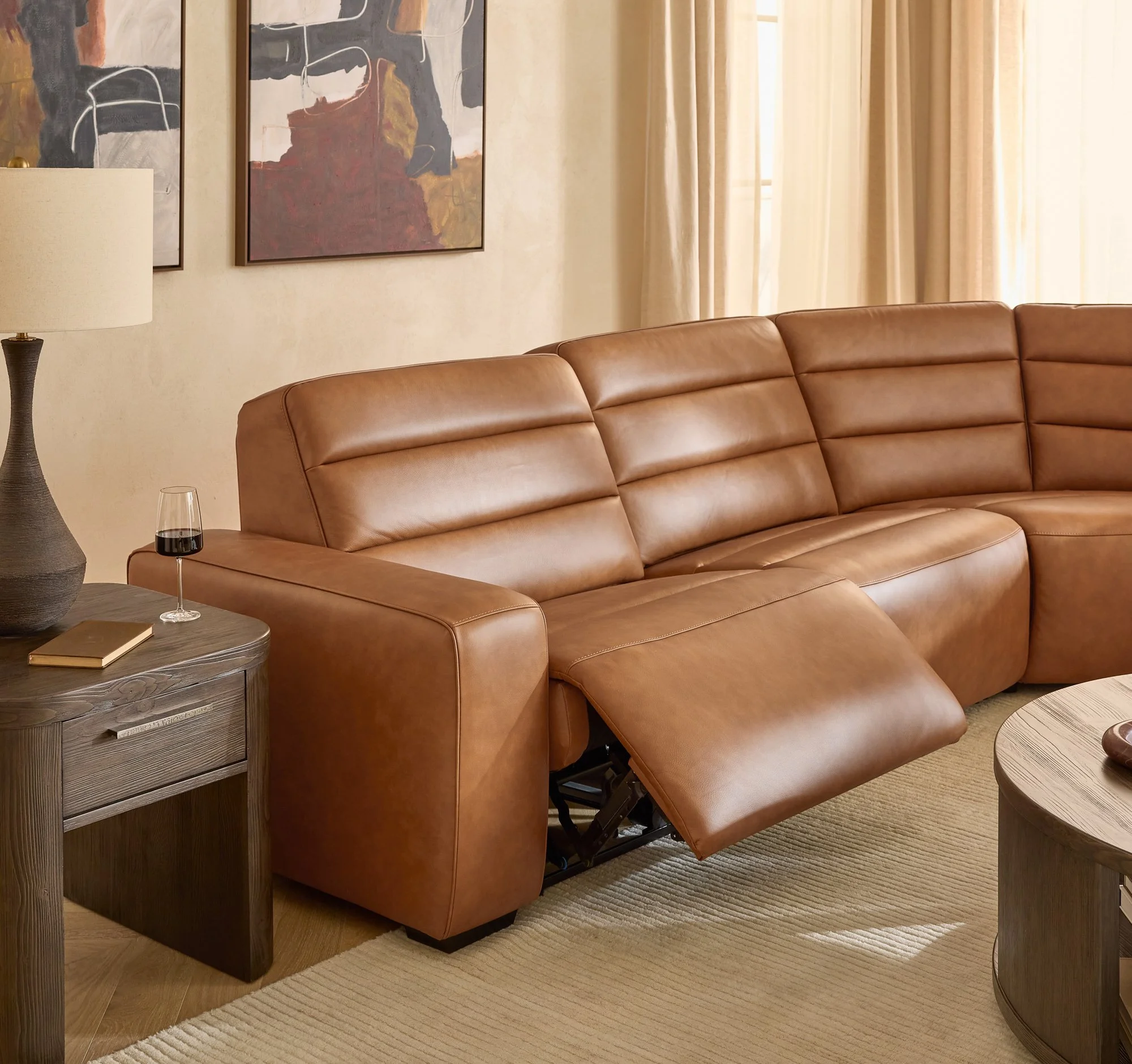 Living room with a tan leather sofa, a wooden side table with a closed book and a glass of red wine, a lamp, and artwork on the wall next to beige curtains.