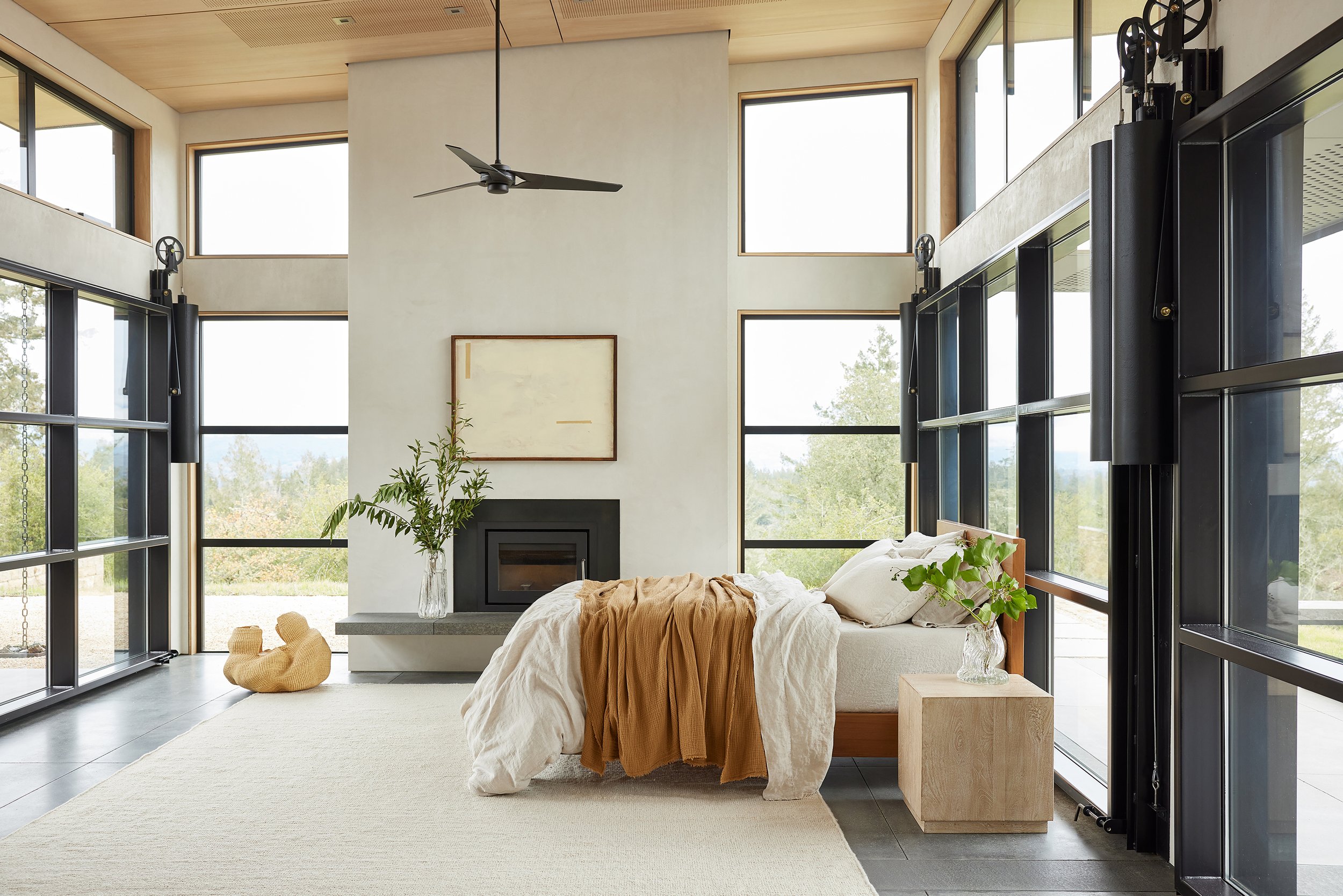 Modern bedroom with large windows, a bed with beige and white linens, a wooden nightstand, a potted plant, and a fireplace.