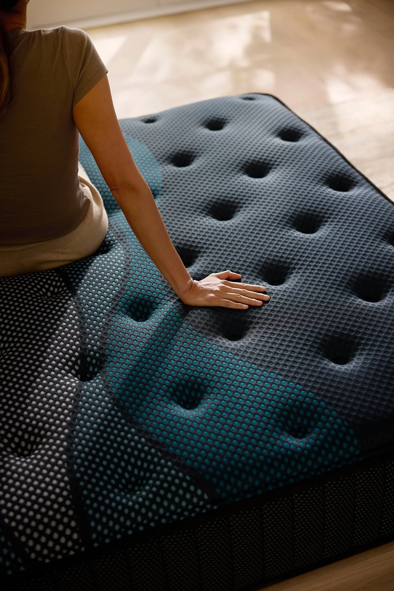 A person kneeling on a black, textured, adjustable air-filled mattress or cushion with large circular holes, on a wooden floor, with sunlight streaming in.