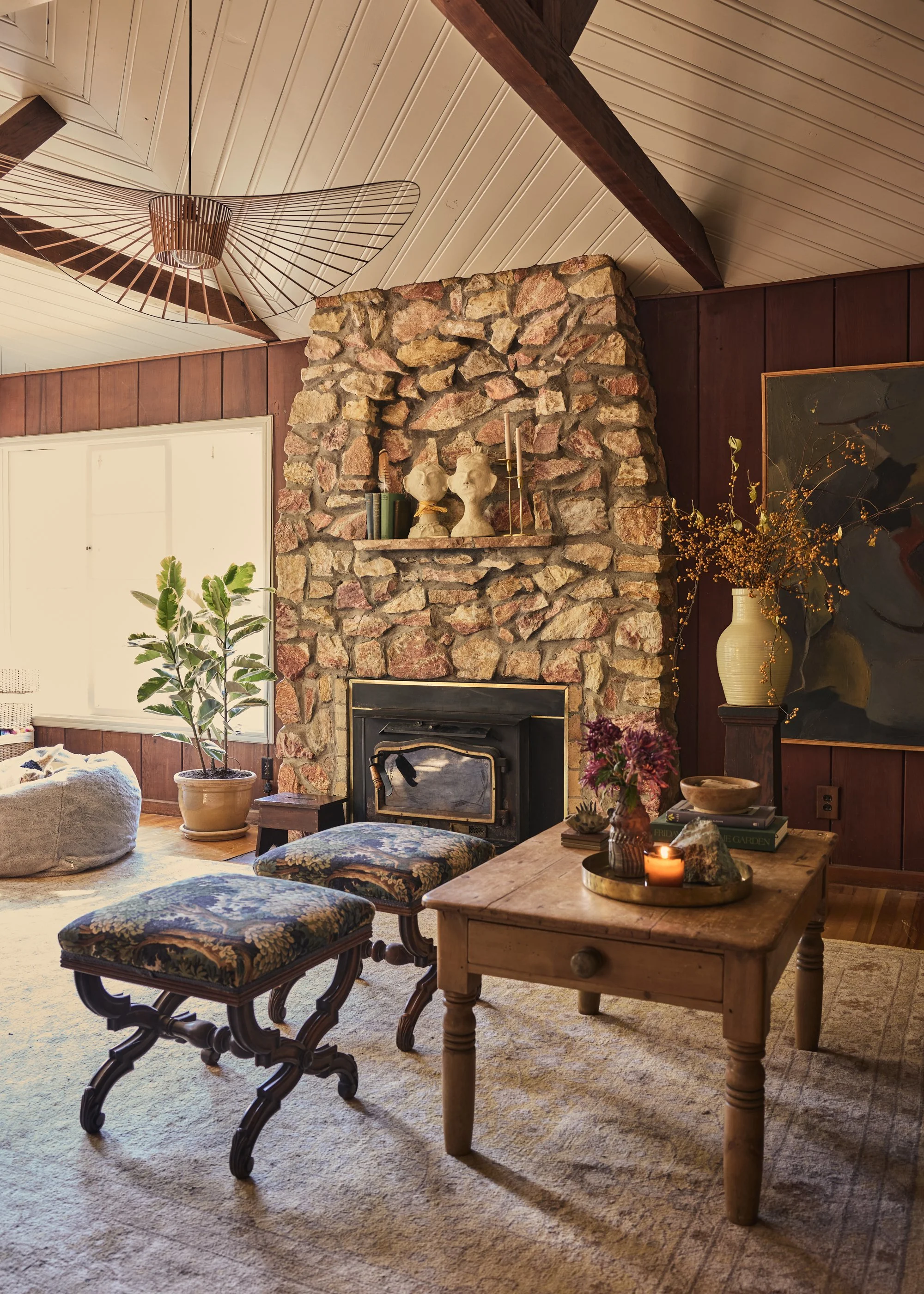 A cozy living room with a stone fireplace, wooden ceiling, and eclectic decor. Features include floral-patterned stools, a wooden coffee table with candles and plants, a large potted plant near a window, and various decorative items on the mantel and