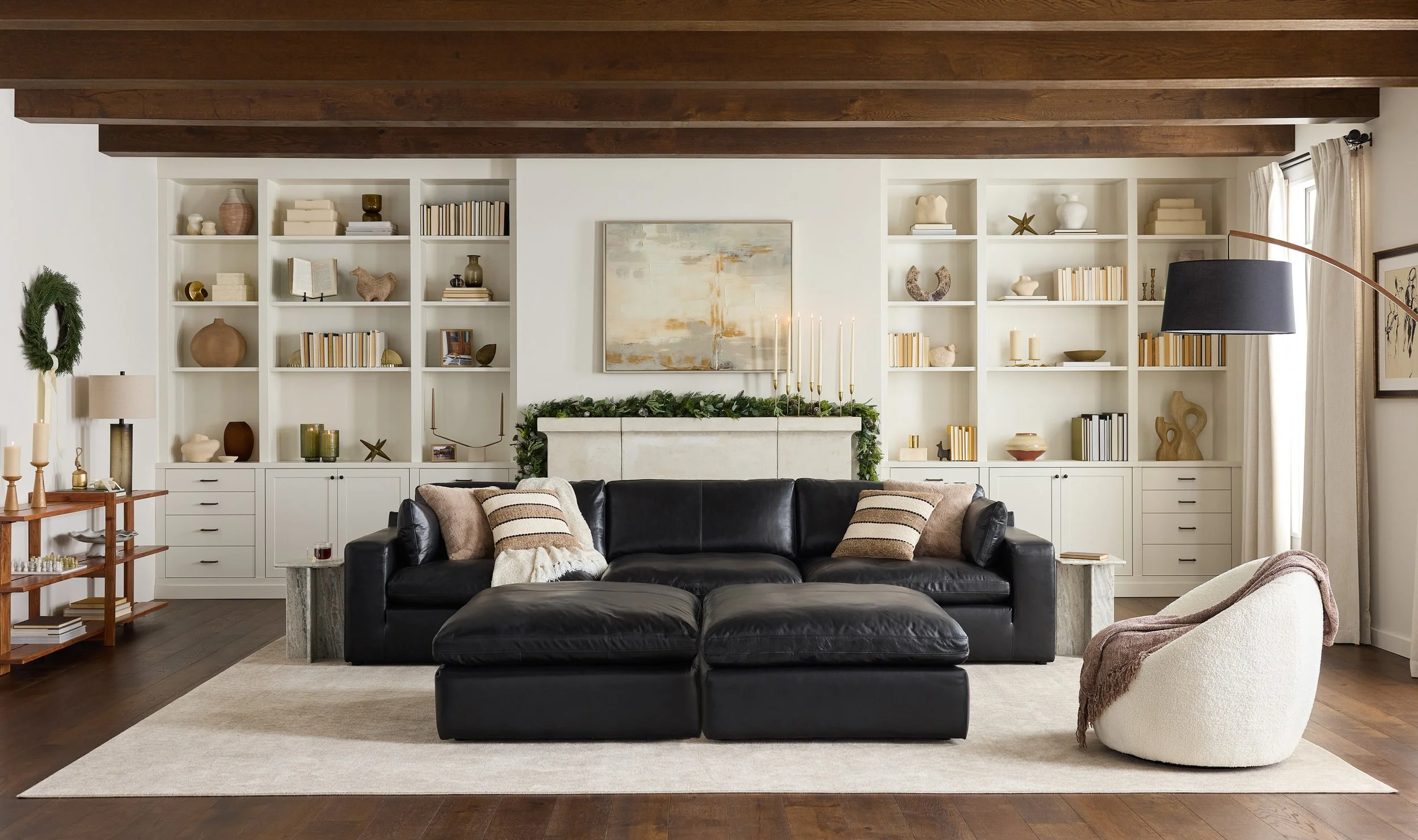 Living room with white built-in bookshelves, beige curtains, a white fireplace decorated with greenery and candles, a black leather sofa with striped cushions, a beige armchair with a throw blanket, and hardwood floors.