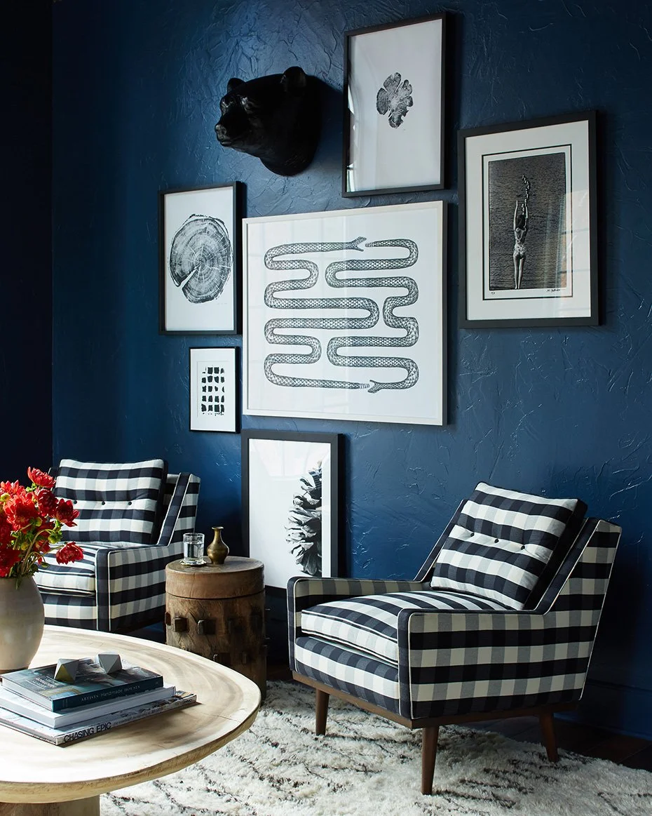 Living room with a deep blue textured wall decorated with framed black-and-white art, a black panther head wall decor, and two black-and-white checkered armchairs. A round wooden side table, a white coffee table with books, and a vase with red flower