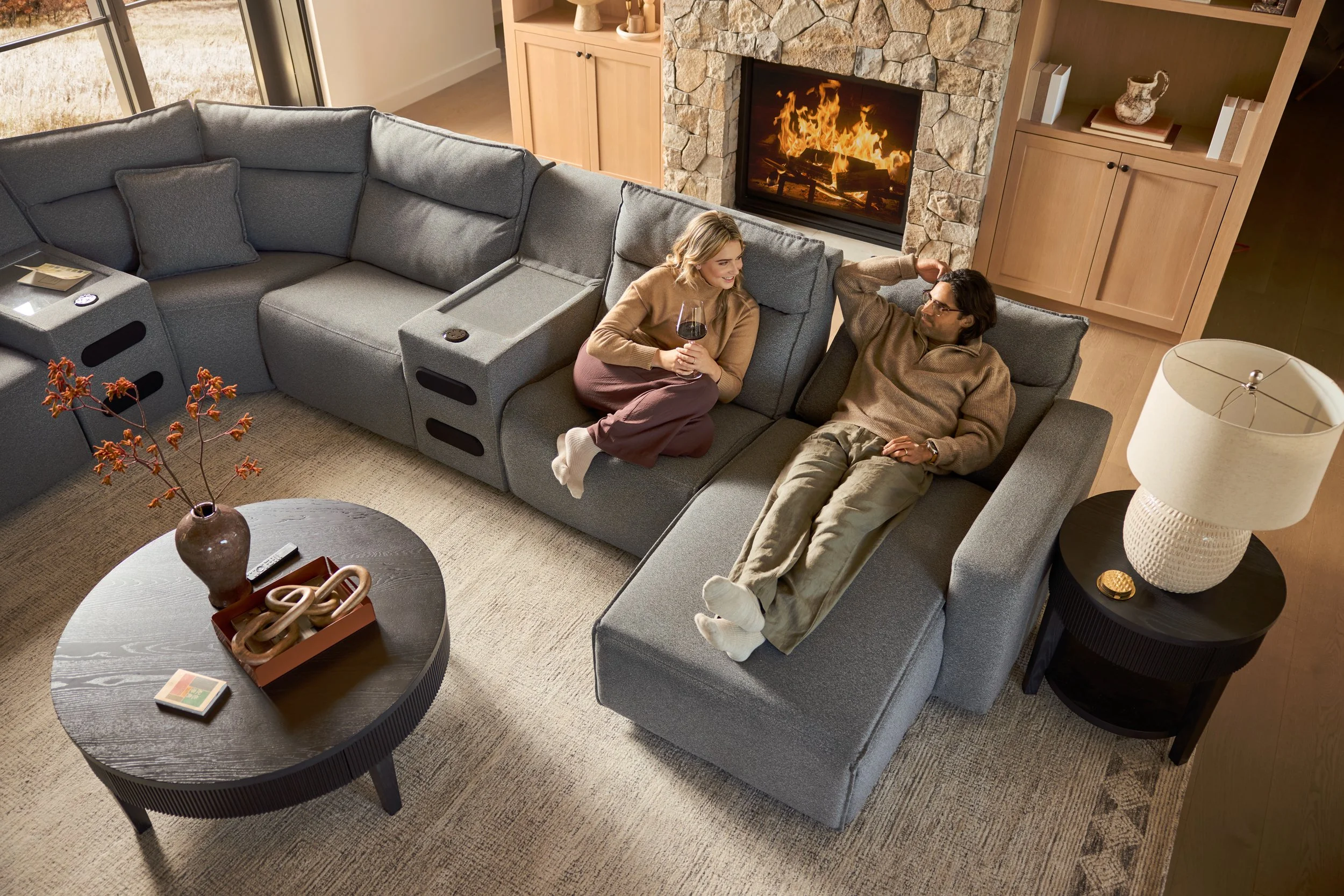 A couple is sitting on a large gray sectional sofa in a living room, with the woman holding a glass of red wine and the man reclining with his arm behind his head. There is a fireplace with a stone surround and a bookcase with decorative items and bo