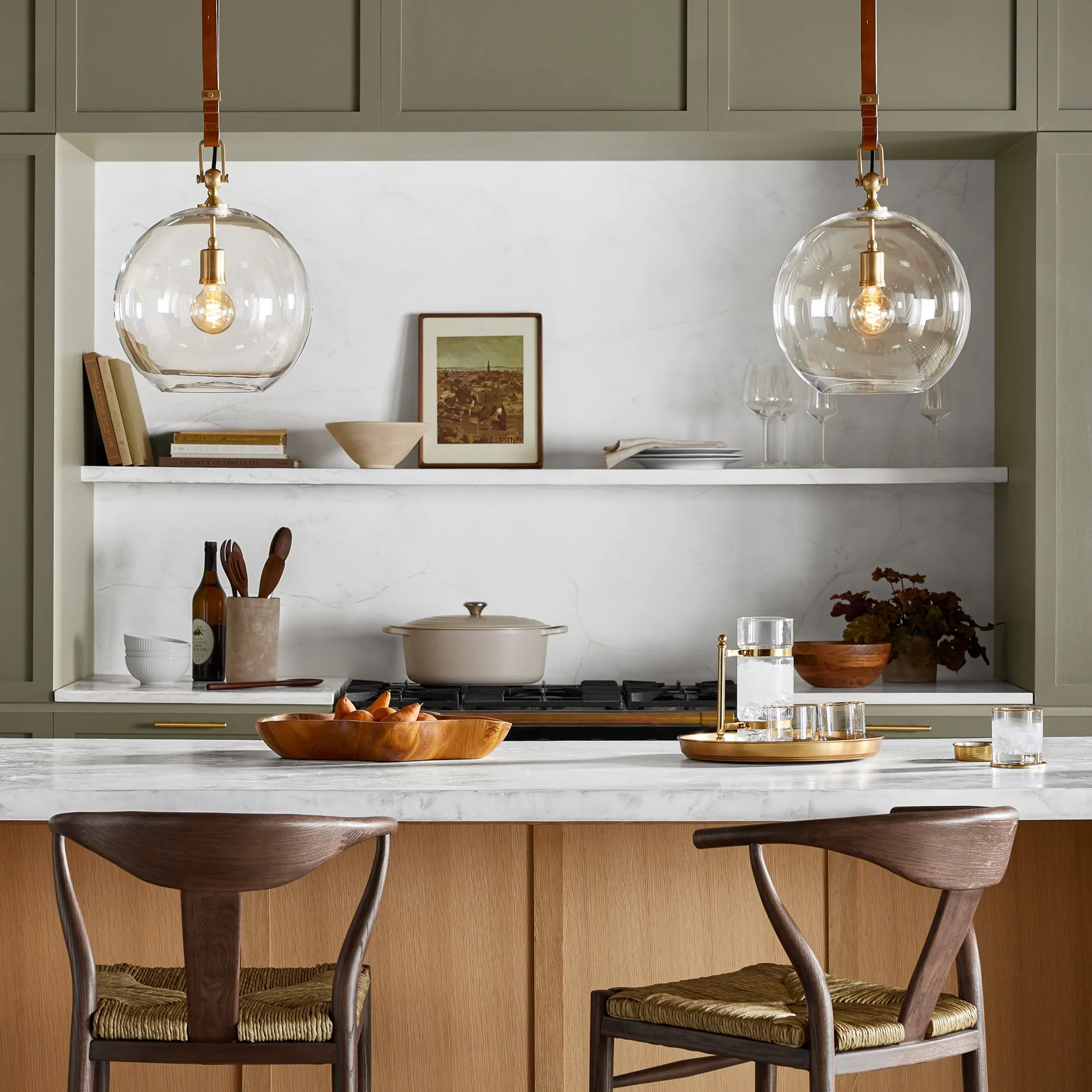 Kitchen with pendant lights, framed artwork, books, bowls, and wine bottles on a white marble countertop, with wooden chairs and cabinetry.