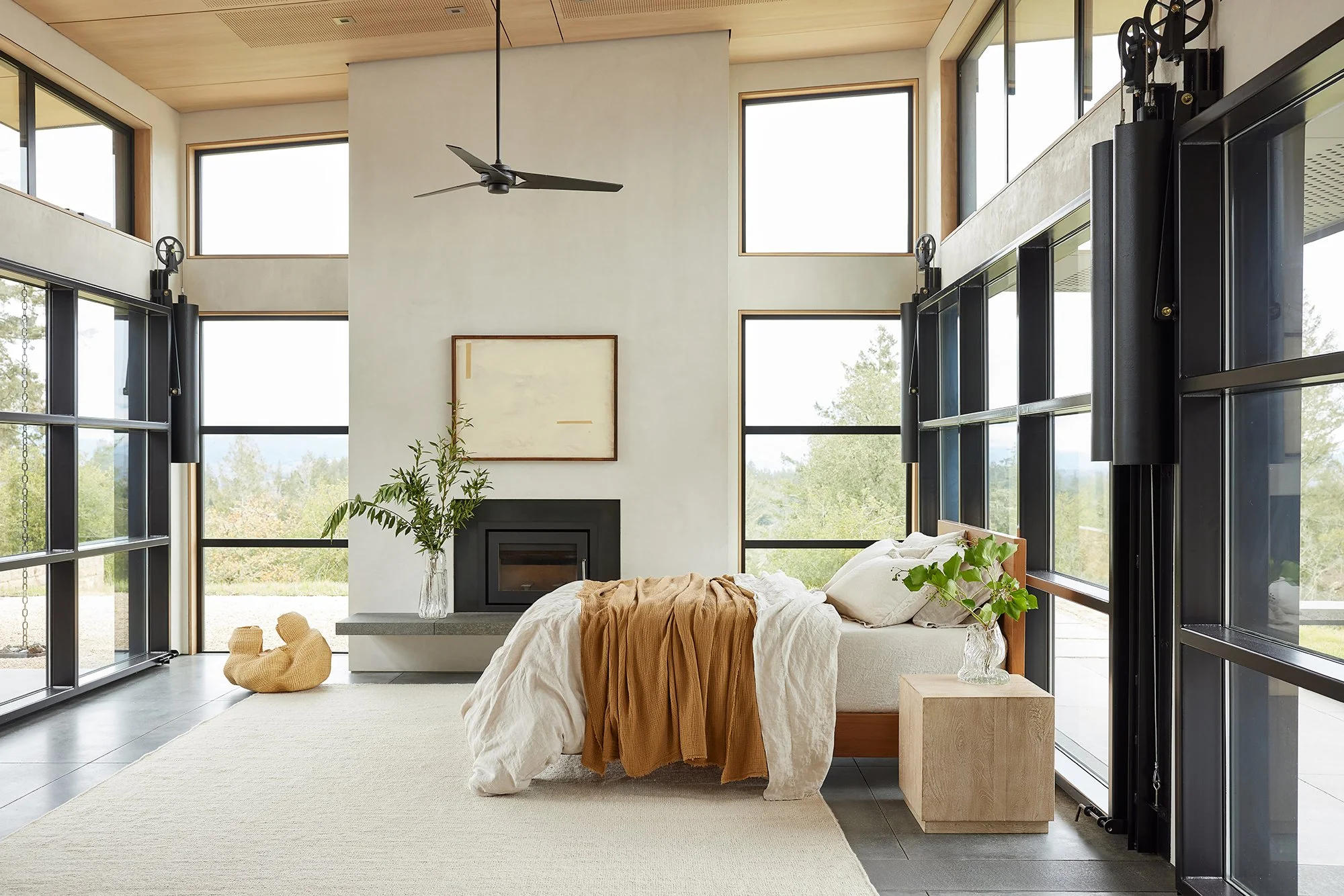 Modern bedroom with large windows, a fireplace, a bed with beige and white linens, a glass vase with green leafy branches, and a wooden side table, with a minimalist and airy design.