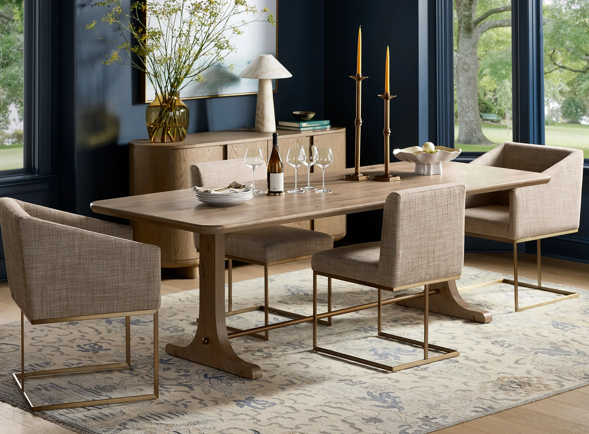 A dining room with a wooden table, upholstered chairs, and a decorative rug. The table has wine glasses, a bottle, a bowl, and candles. A sideboard with a vase of flowers, a lamp, and stacked books is visible. Large windows reveal a green outdoor sce