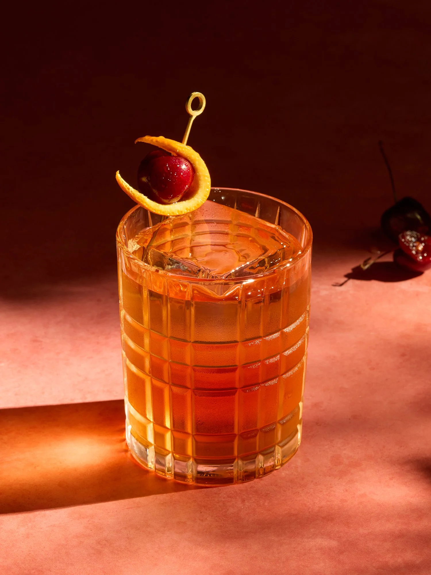 A glass of orange-colored cocktail with a cherry and orange twist garnish, set on a pinkish surface with a dark background.