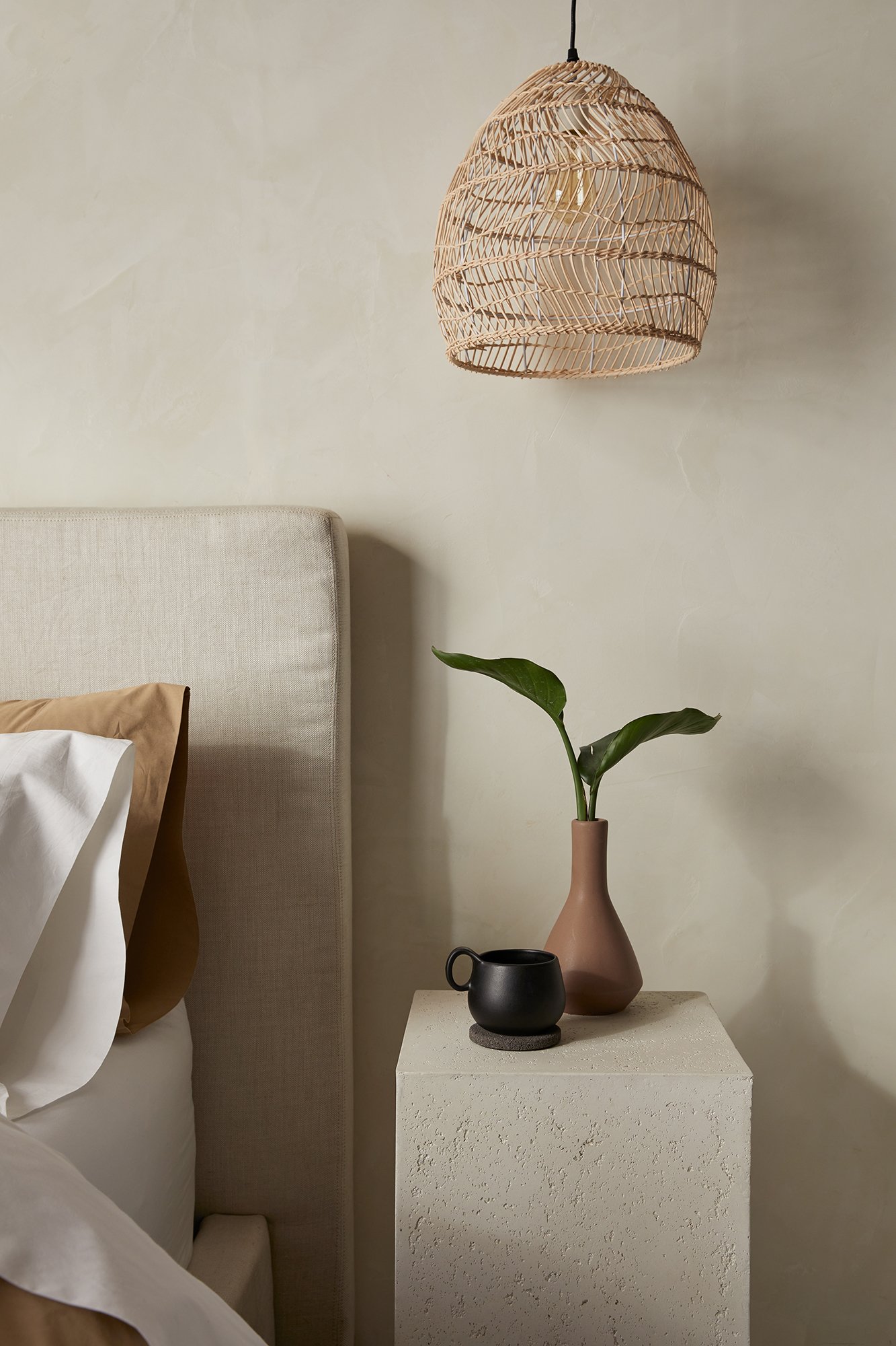 A bedroom scene featuring a wall-mounted wicker pendant light, a beige upholstered headboard, a bed with white sheets and brown and white pillows, a beige vase with a green leafy plant, and two small black and gray cups on a textured white bedside ta