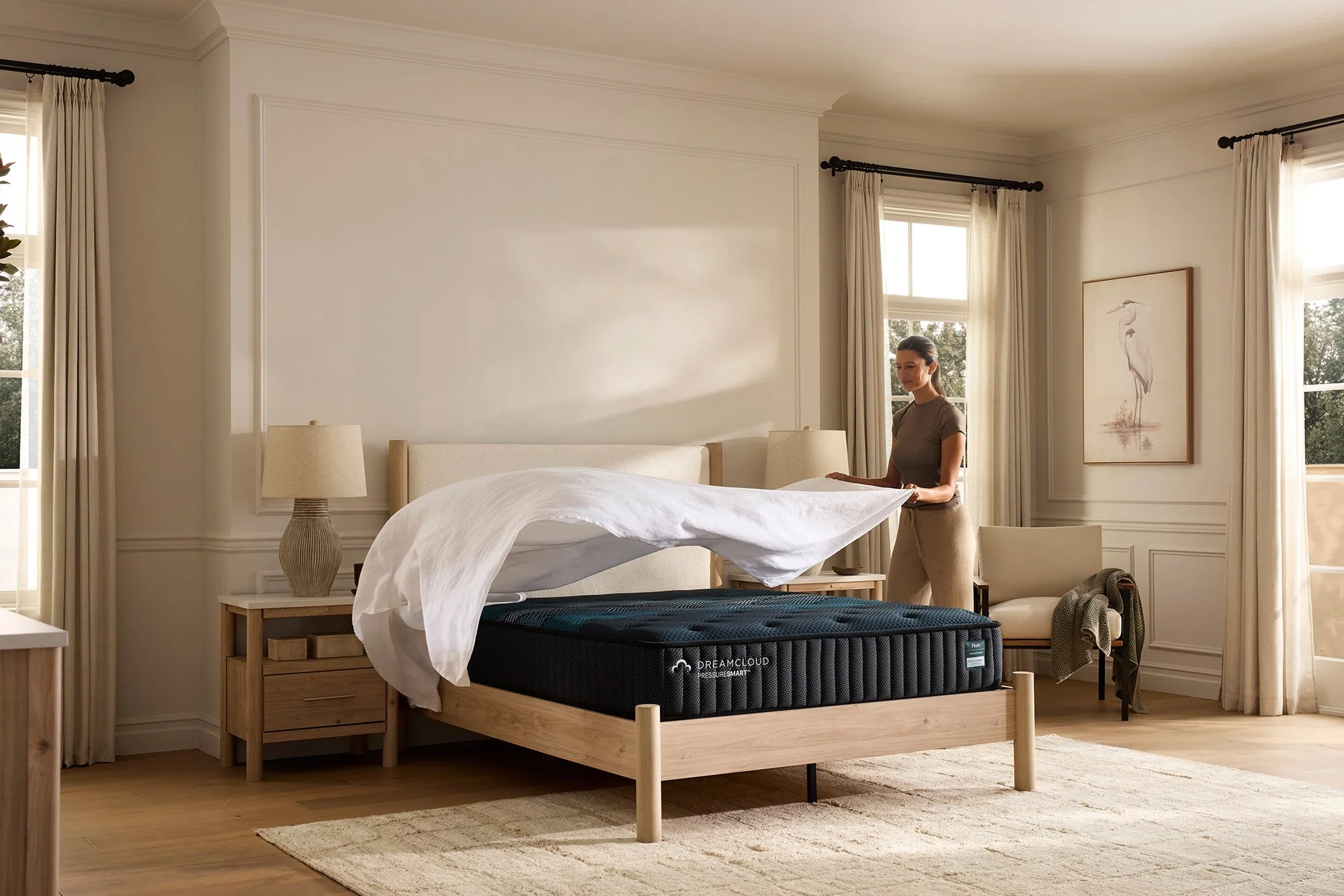 A woman making a bed with a mattress in a well-lit modern bedroom with large windows, beige curtains, framed artwork of a heron, and wooden furniture.
