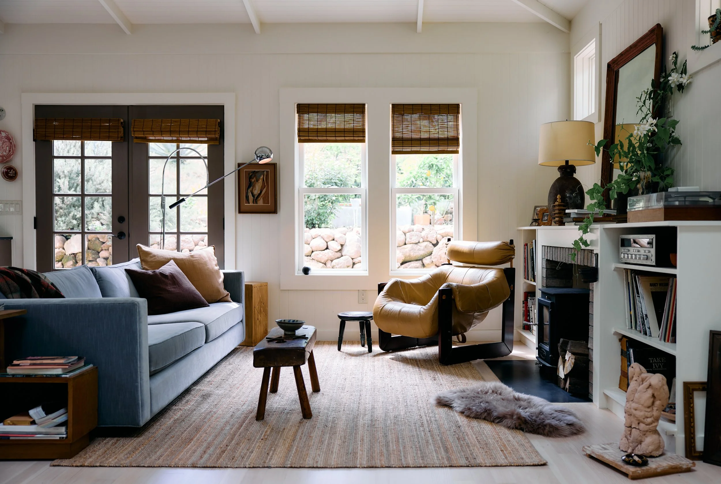 Living room with a blue sofa, tan armchair, bookshelf, fireplace, and large windows with bamboo blinds, decorated with artwork and plants.