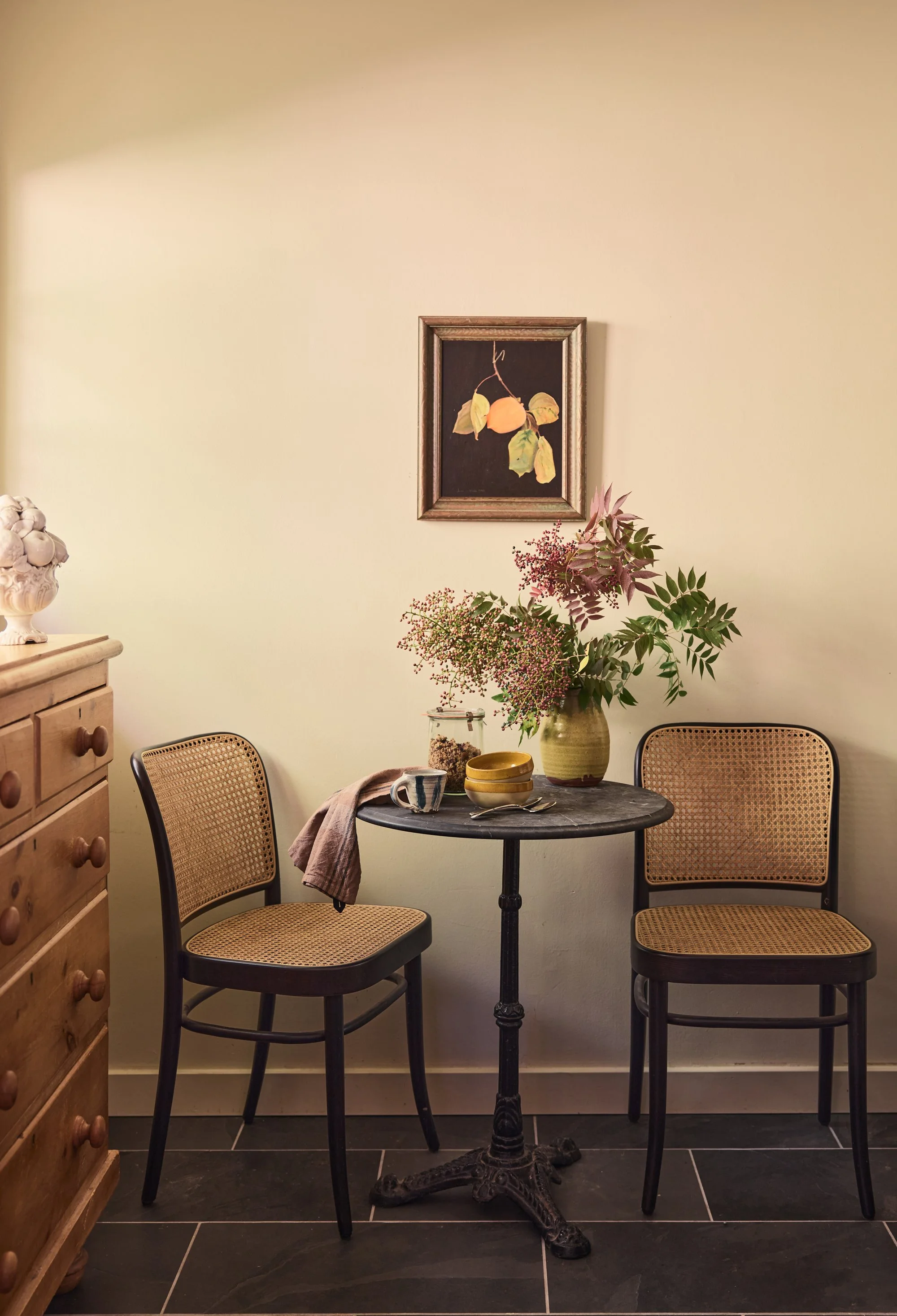 A small round black table with two cane-backed chairs, a large green and pink vase with flowers, and some bowls and teacups on the table. There is a wooden dresser on the left and a framed painting of a lemon with leaves hanging on the cream-colored 